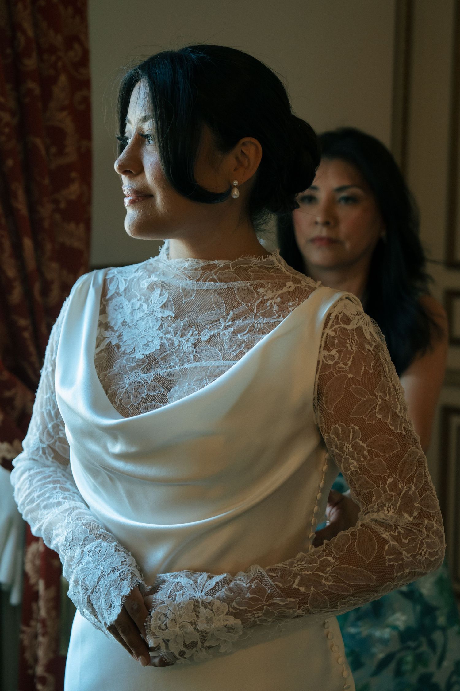Bride in elegant white lace long-sleeve wedding gown with draped back, standing with bridesmaid in softly lit room.