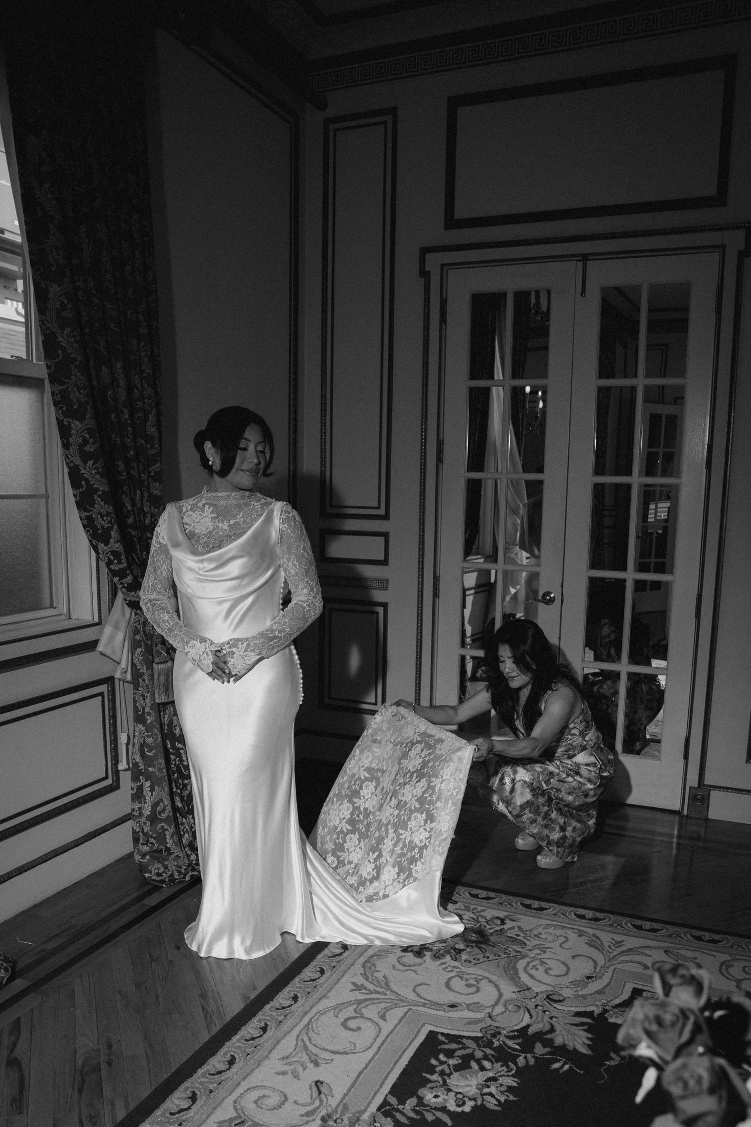 Black and white photo of a bride in a lace wedding gown being assisted by a woman adjusting her train in an elegant room.