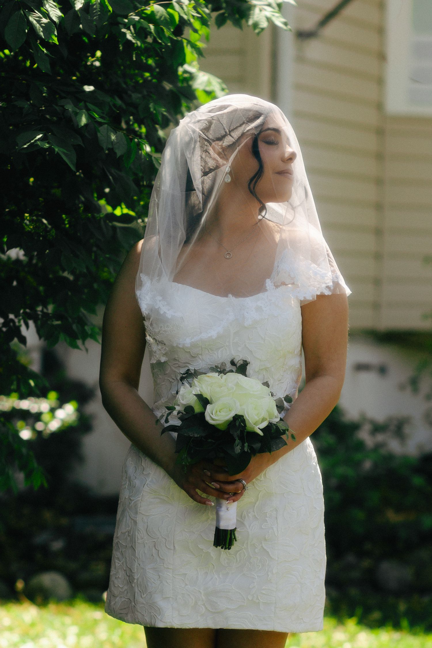 Bride in short white lace dress and veil holds white rose bouquet outdoors near green trees.