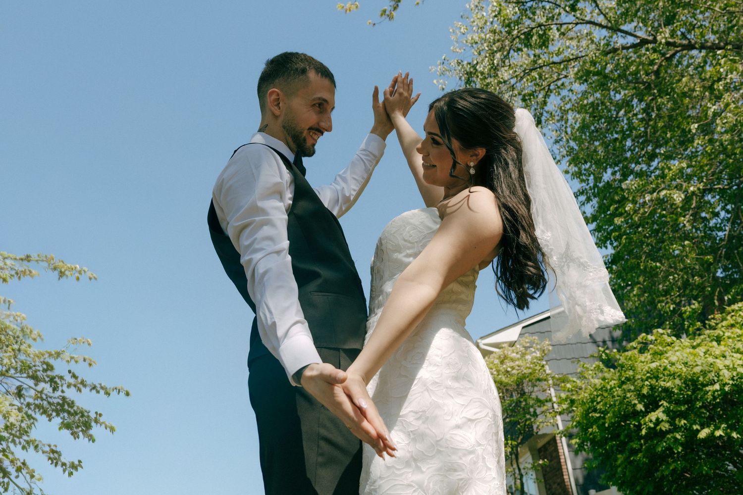 Bride and groom dancing outdoors on sunny day, bride in white gown with veil, groom in black vest, green trees behind.