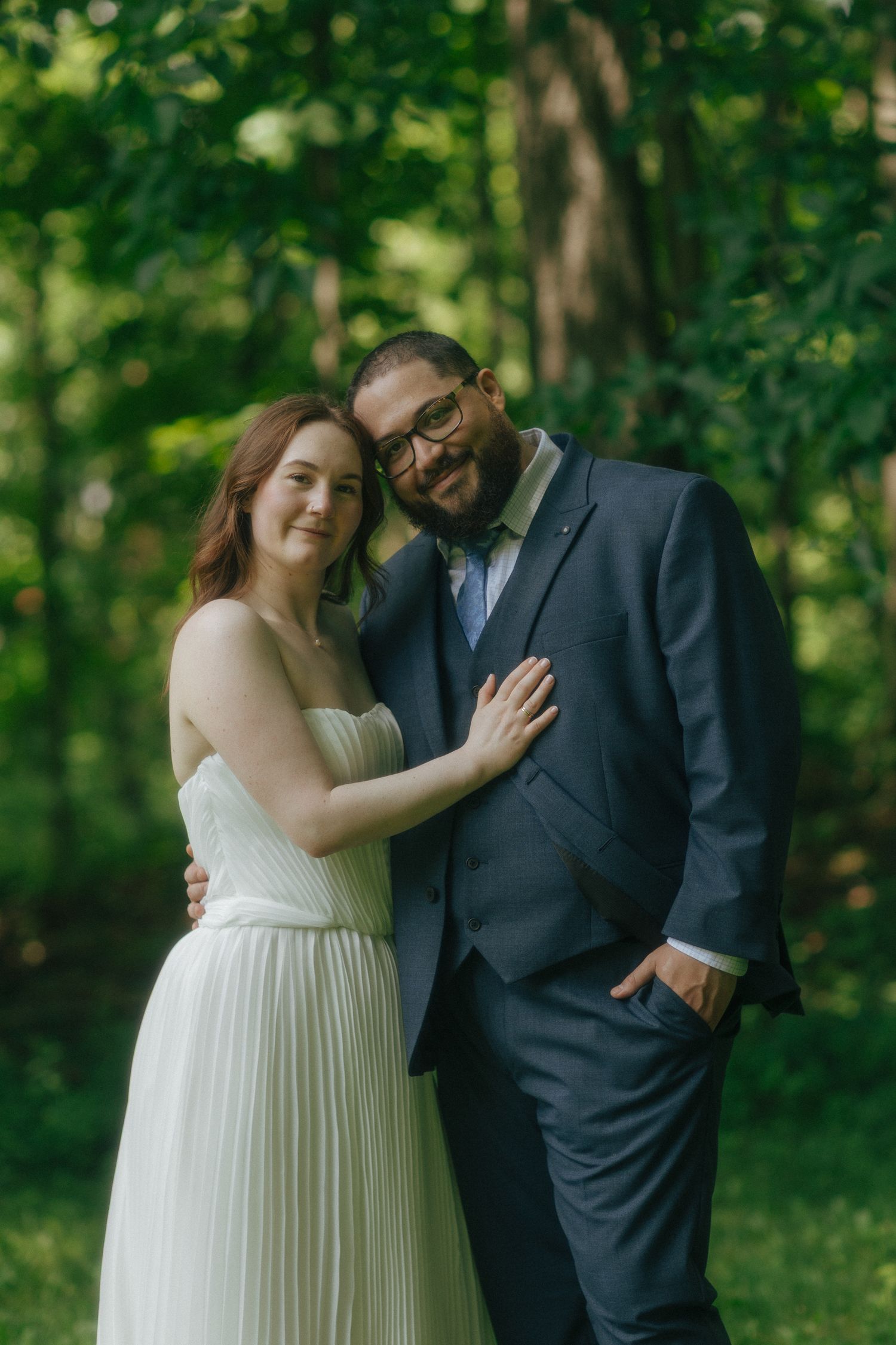 A couple in wedding attire poses outdoors in a lush green forest setting, smiling warmly together.