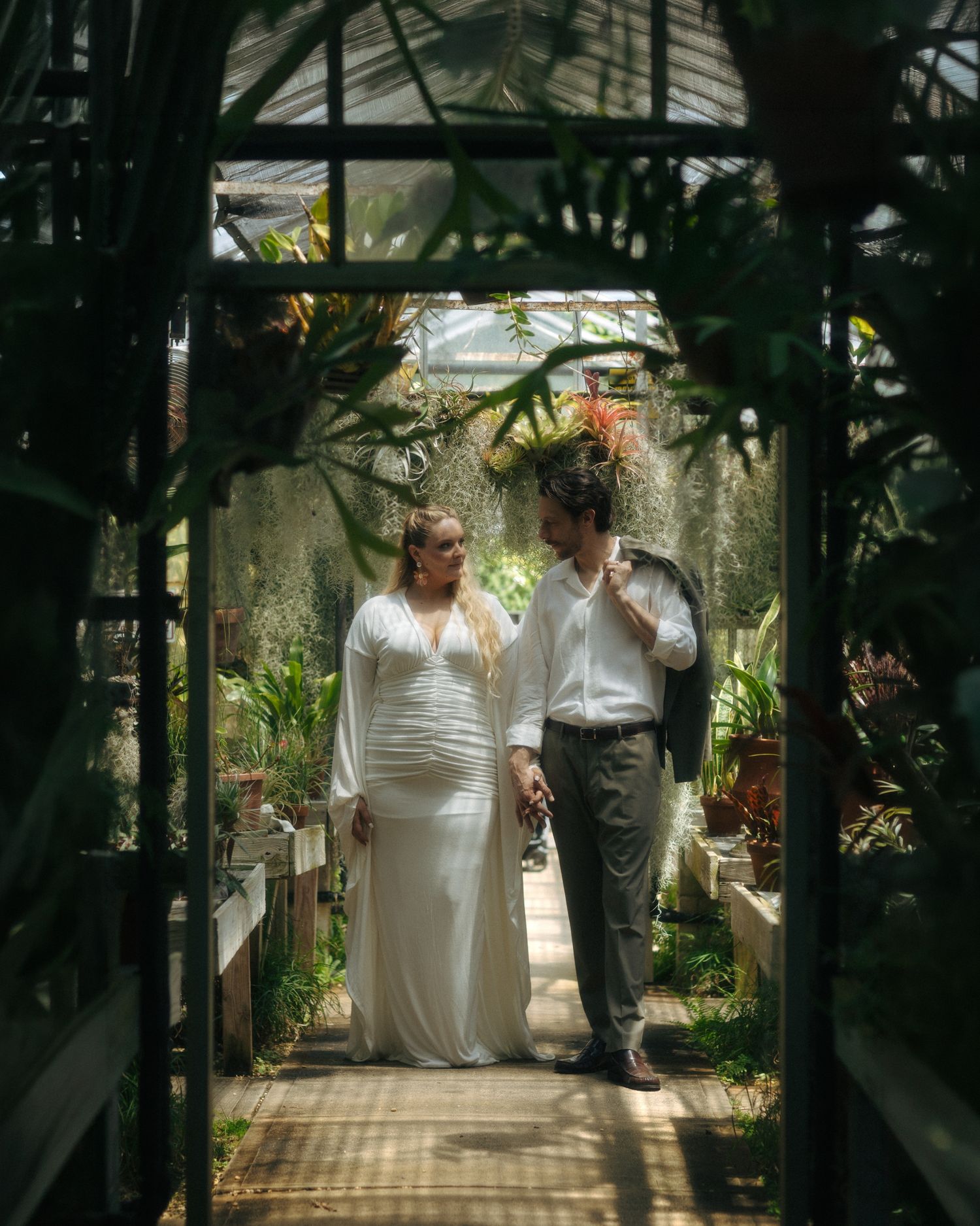 A couple in wedding attire shares a romantic moment inside a lush greenhouse surrounded by tropical plants.