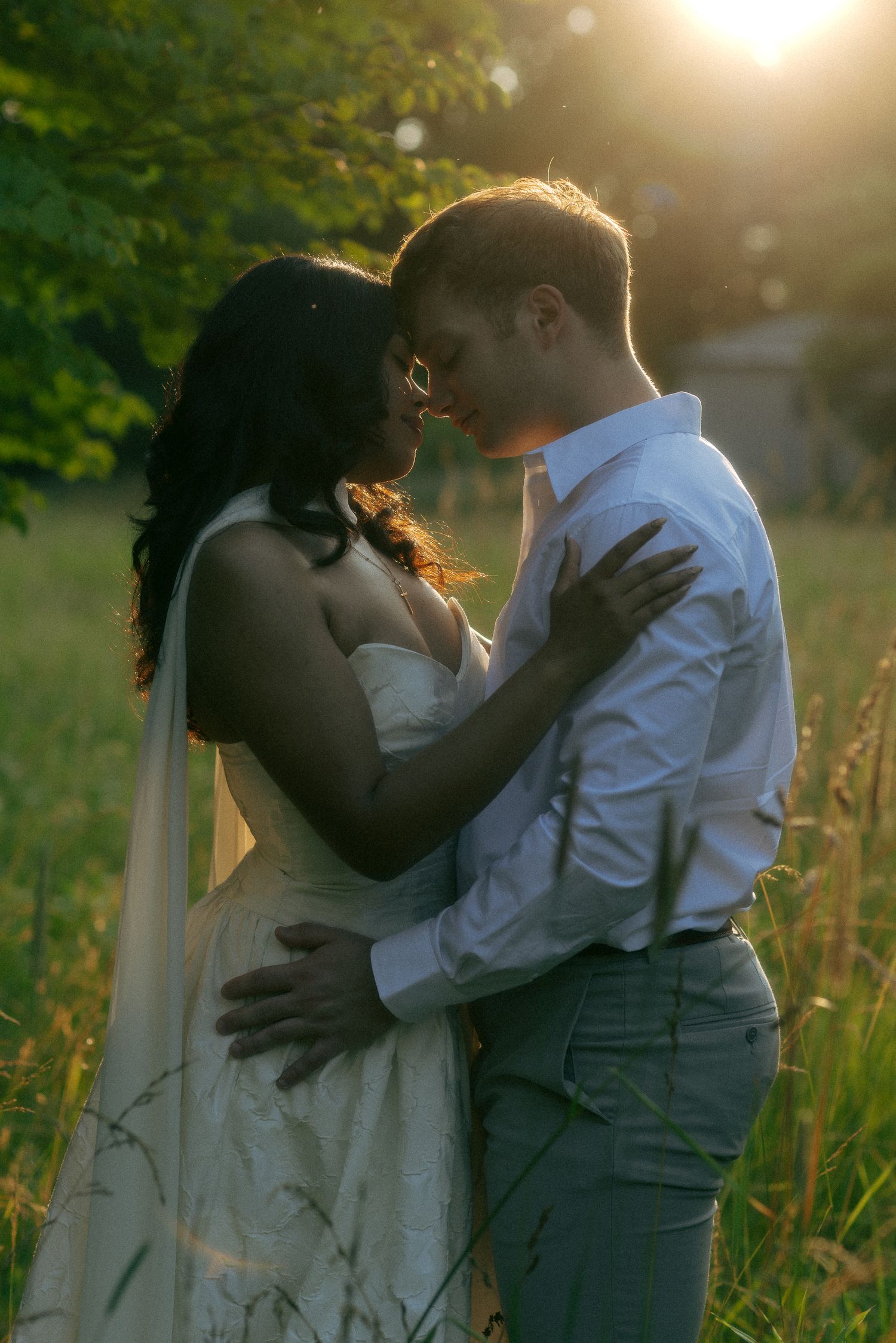 Romantic couple embracing in golden sunset light in a lush green field, bride in white gown with veil.
