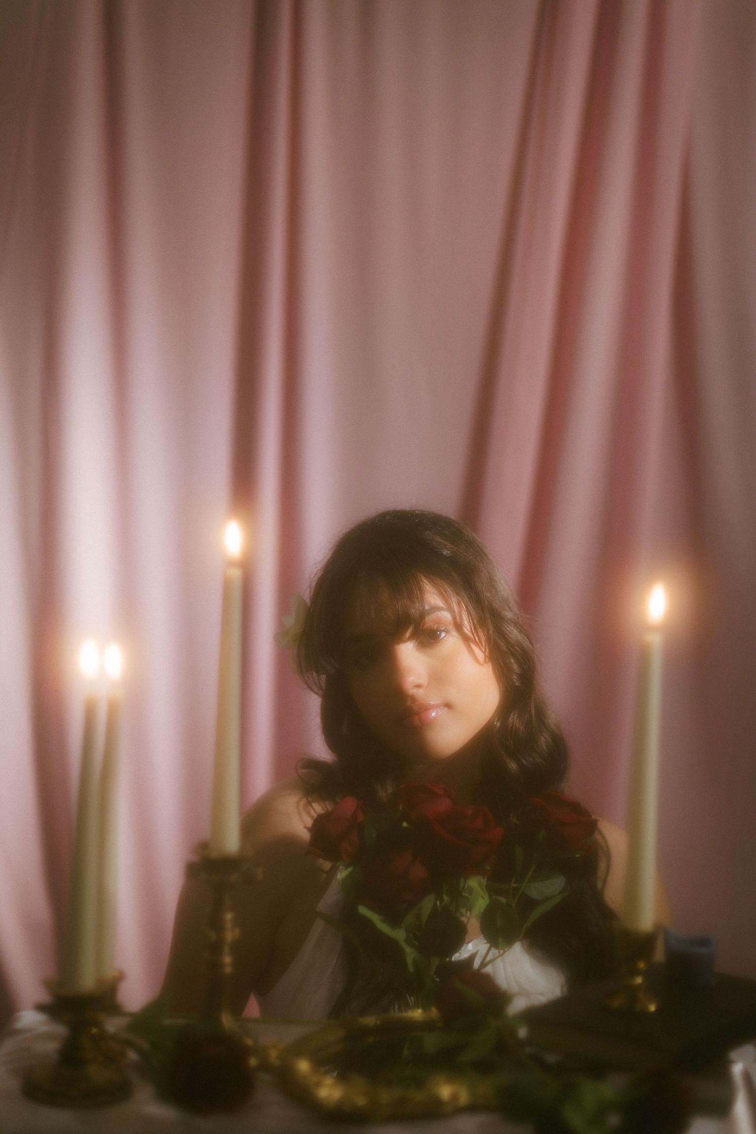 Moody portrait of woman with red roses at candlelit table with pink curtain in background.