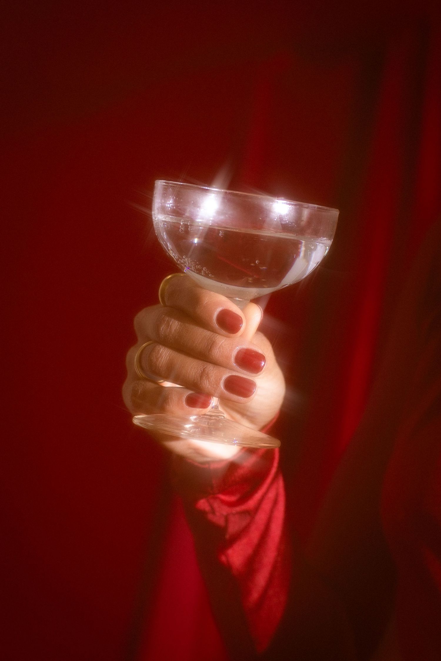 Close-up of hand with red nails holding glowing crystal martini glass against red fabric.