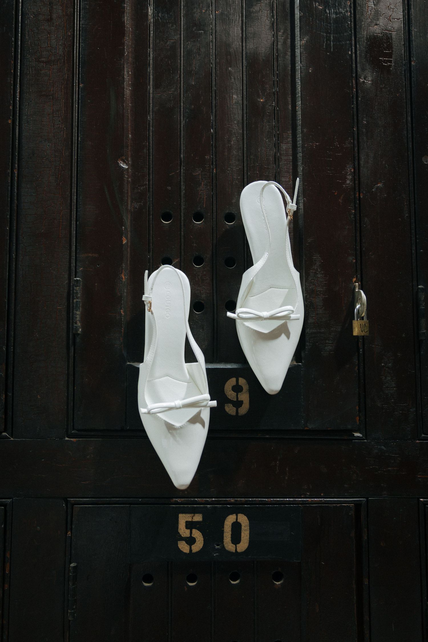 White bridal heels with bow detail hanging on dark wooden locker number 50.
