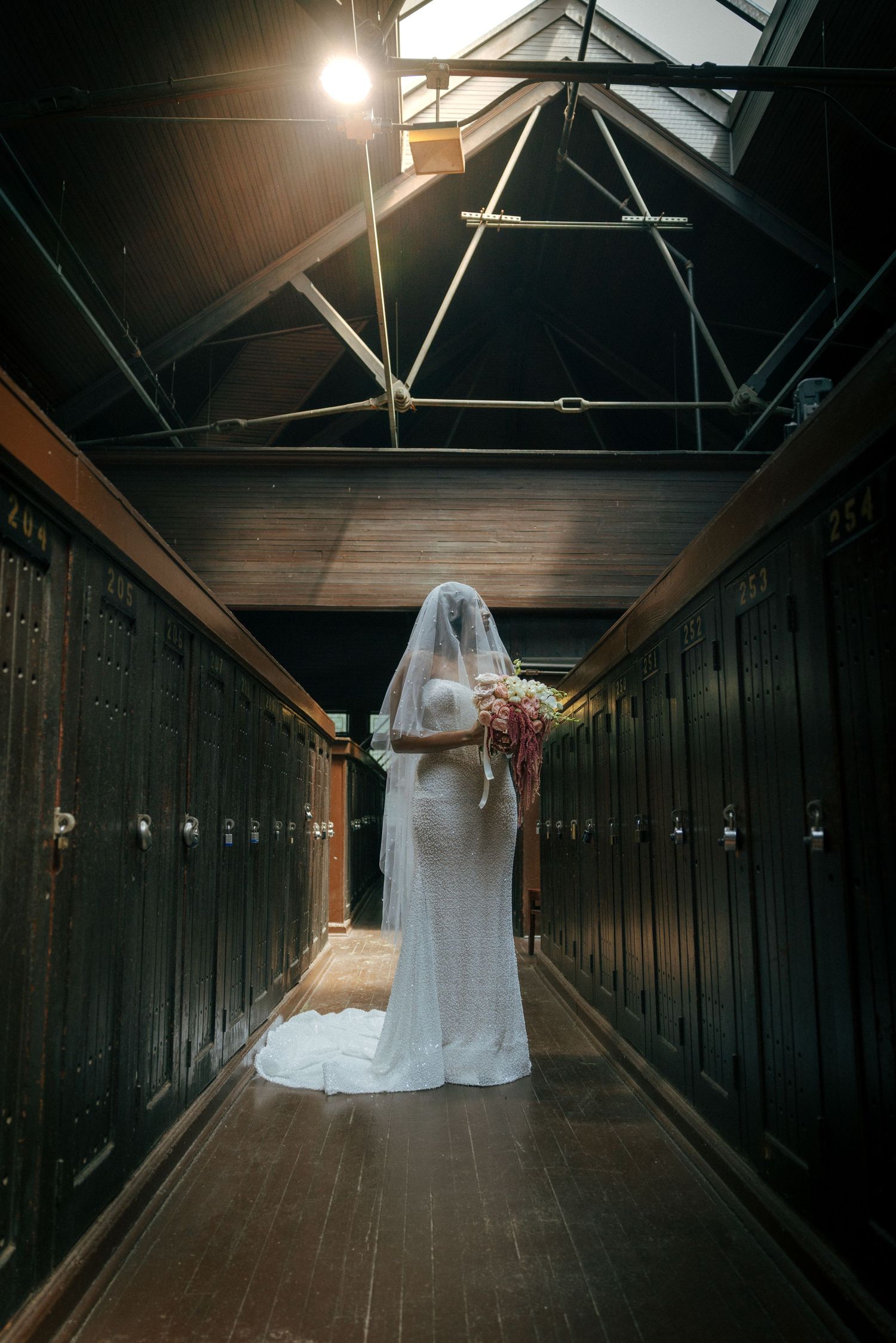 Bride in white gown and veil holding bouquet stands in dimly lit locker room hallway.