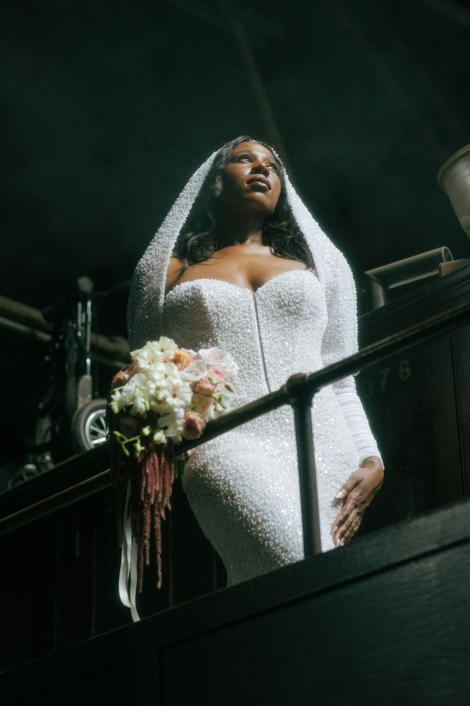 Bride in pearl beaded gown and veil holding bouquet on dark venue balcony.