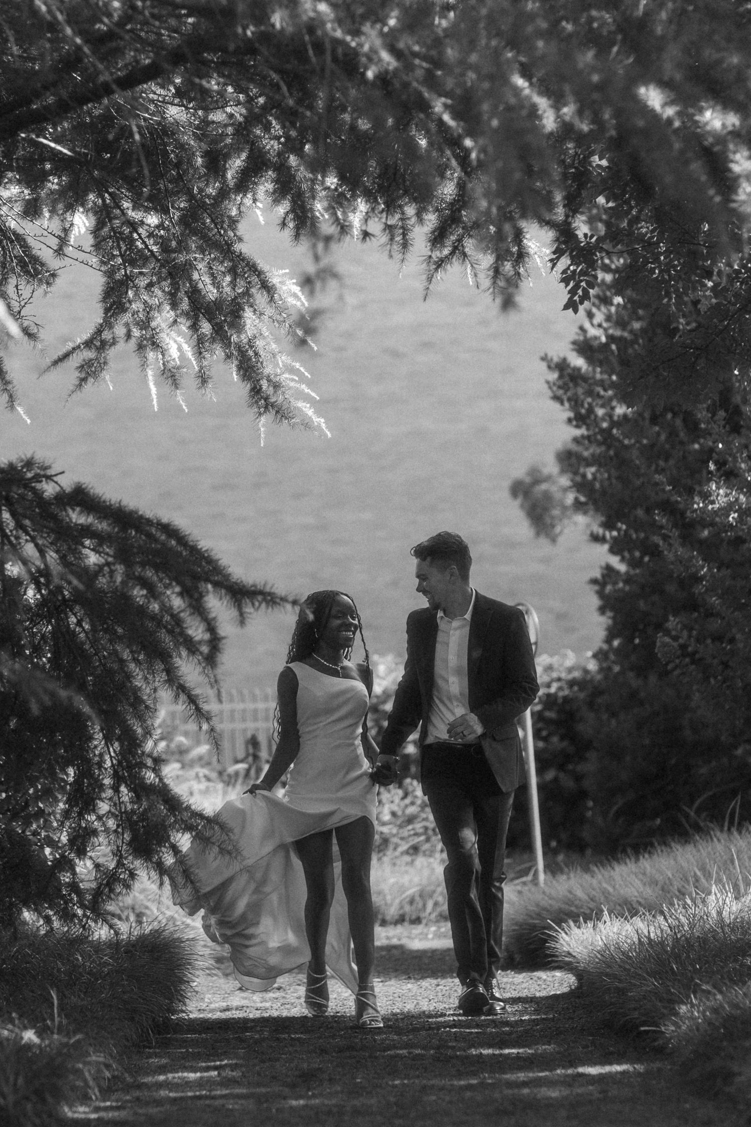 Black and white photo of a couple in wedding attire walking hand in hand along a tree-lined garden path.