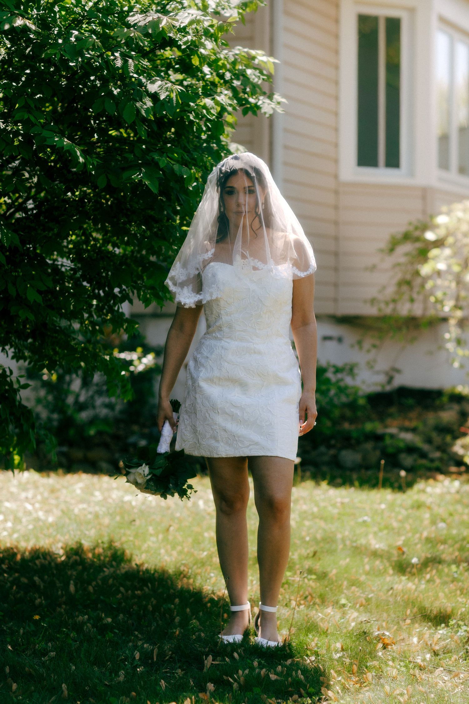 Bride in short white lace dress and veil stands on green lawn beside flowering shrub near house.