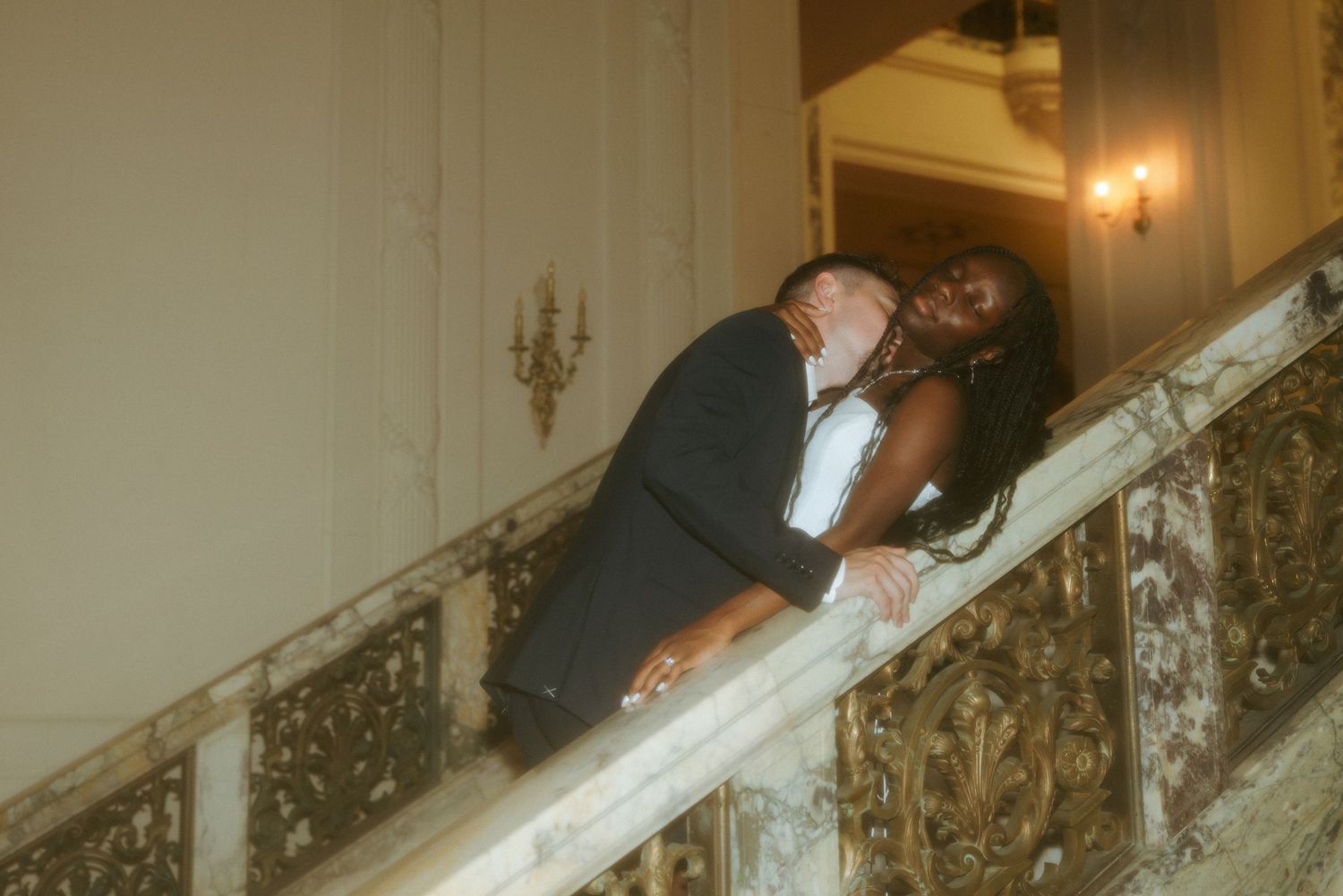 Couple sharing a romantic embrace on an ornate marble staircase, groom in tuxedo and bride in white gown.
