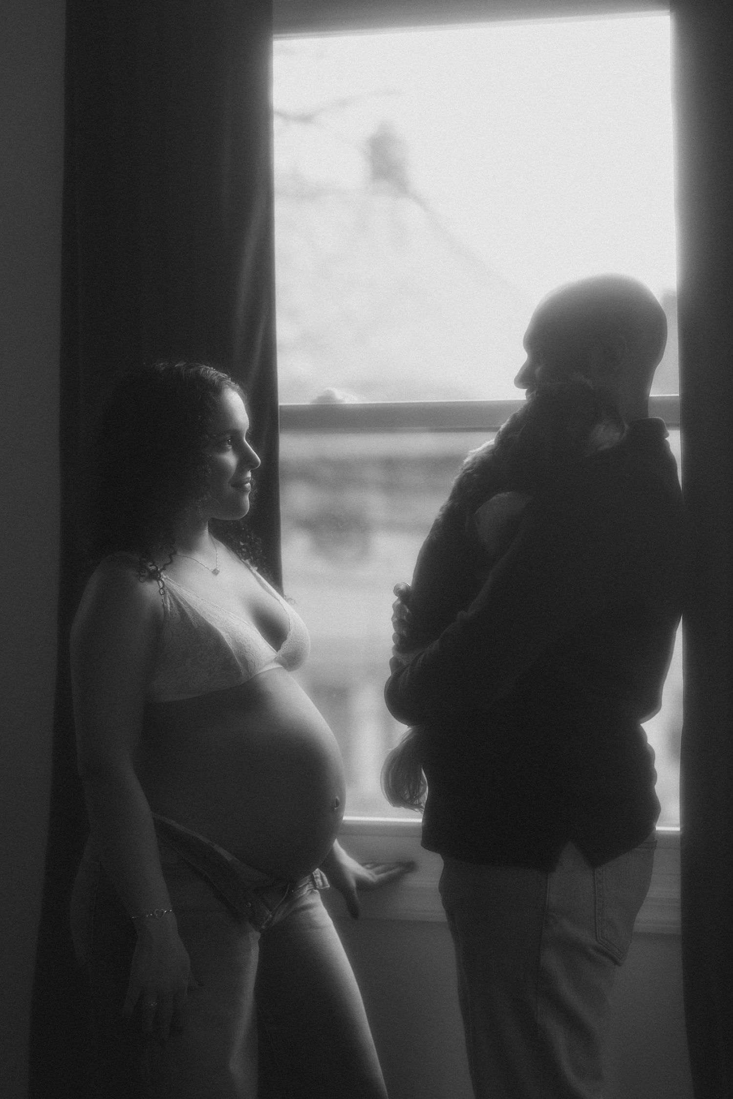 Black and white maternity photo of pregnant woman in lingerie standing by window with partner.