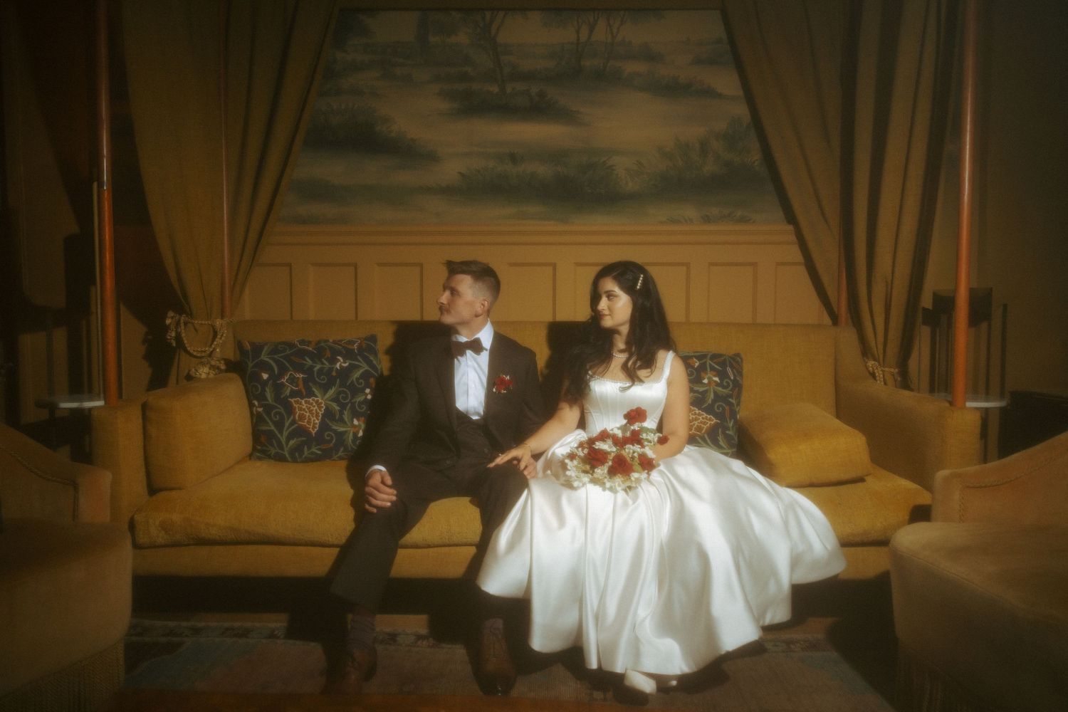 Bride and groom sitting together on an elegant gold sofa, posing for a classic wedding portrait indoors.