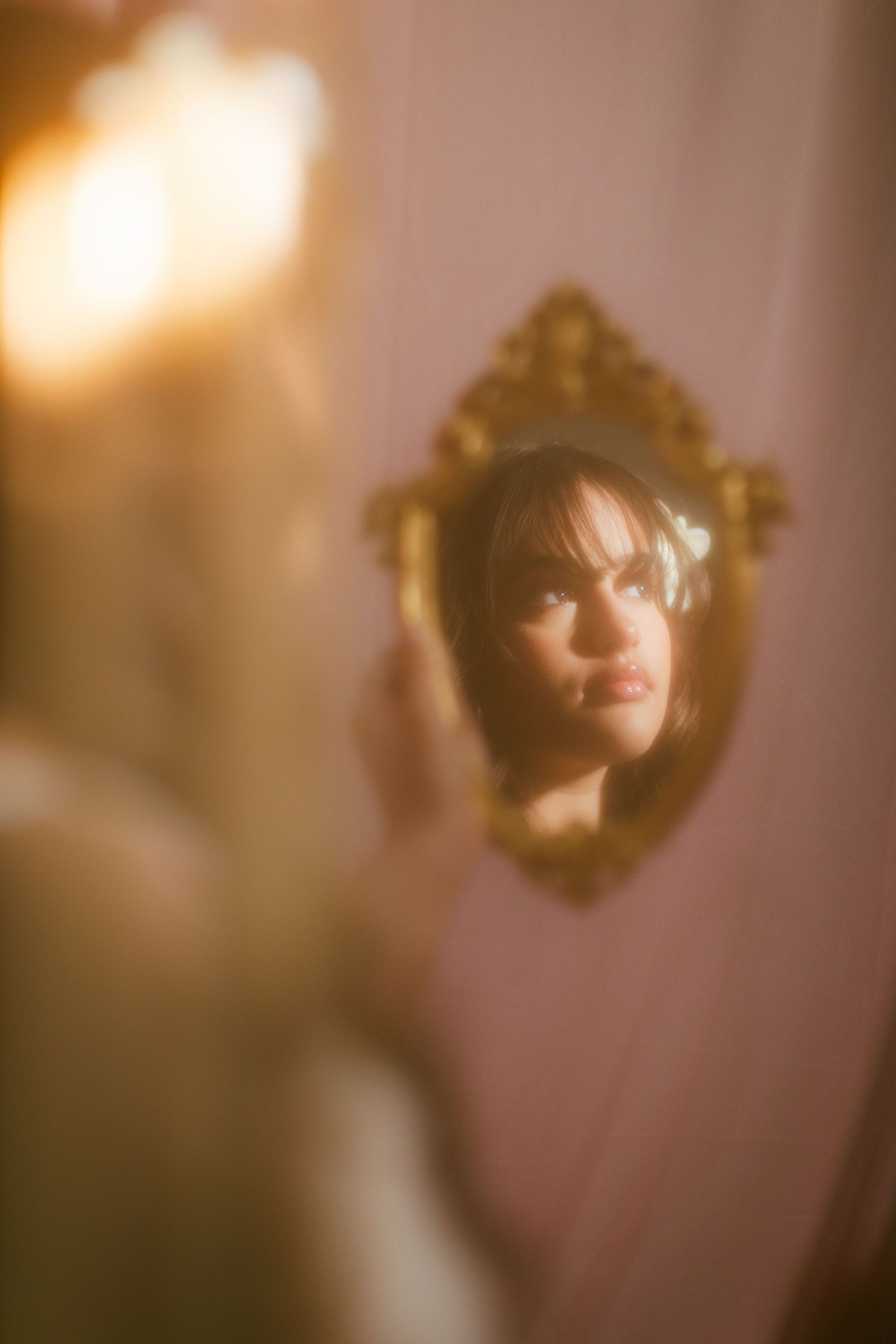Woman's reflection in ornate gold mirror against soft pink blurred background.