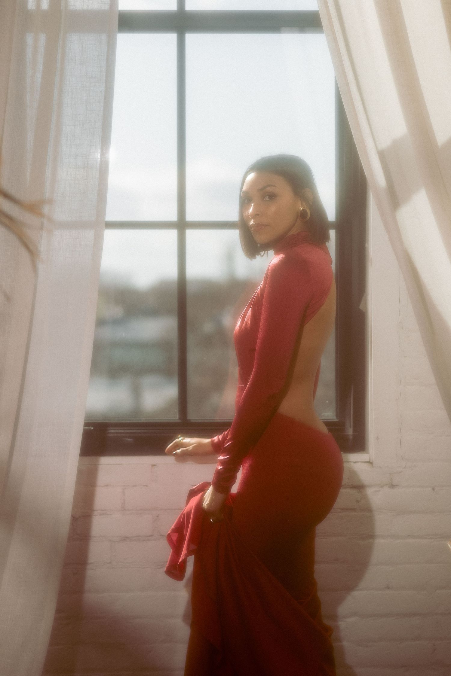 Woman in backless red gown posing by large window with soft natural light filtering in.