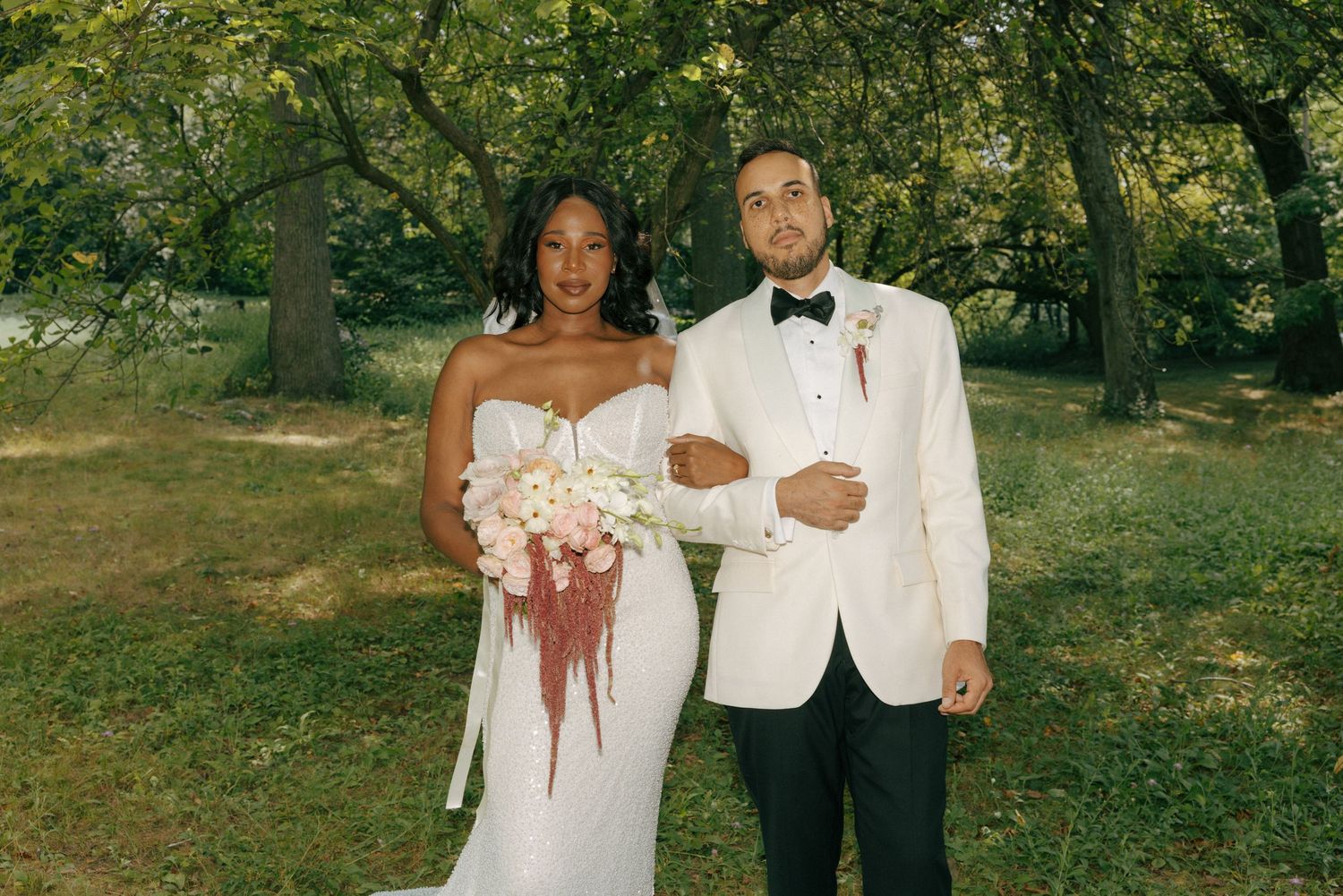 Wedding couple posing in white formal attire with pink bouquet in green park.