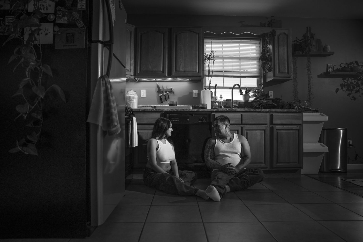 Black and white photo of two people sitting on kitchen floor, sharing an intimate conversation at home.