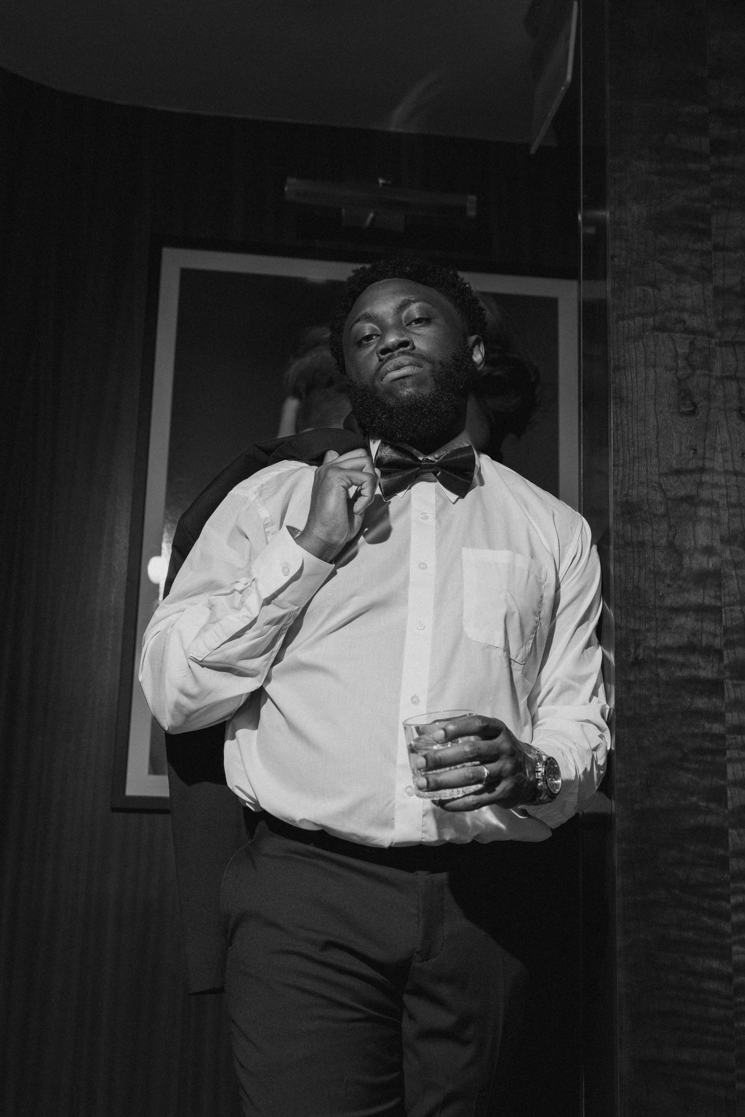 Black and white photo of a well-dressed man in a white shirt and bow tie holding a drink indoors.