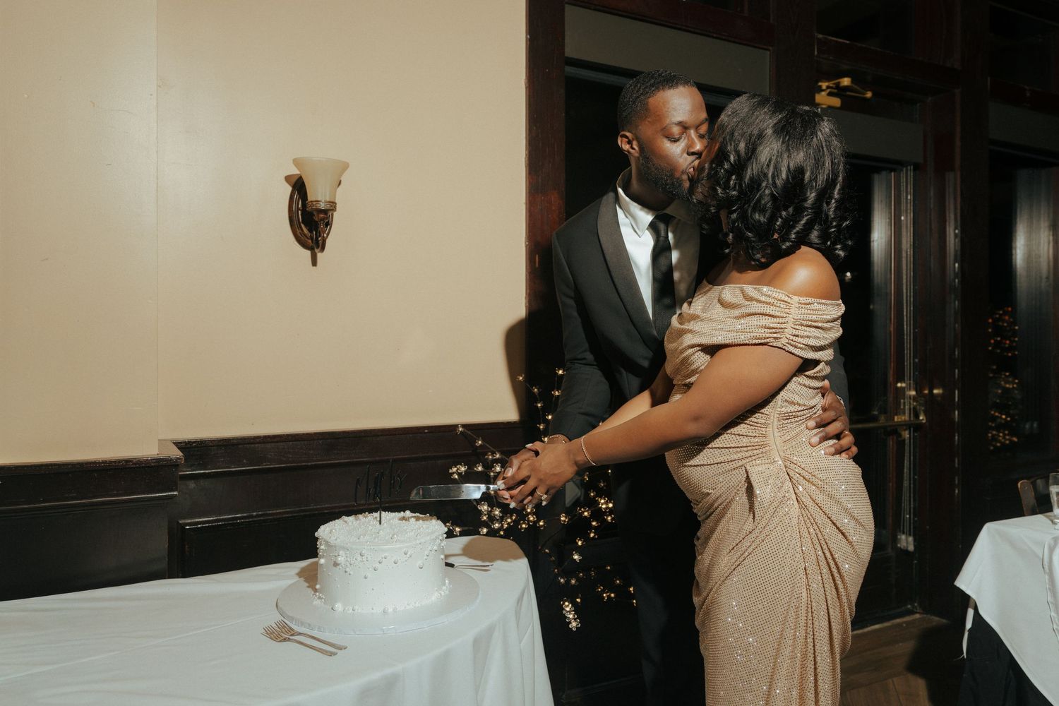 A couple shares a kiss while cutting their white wedding cake at a elegant reception venue.