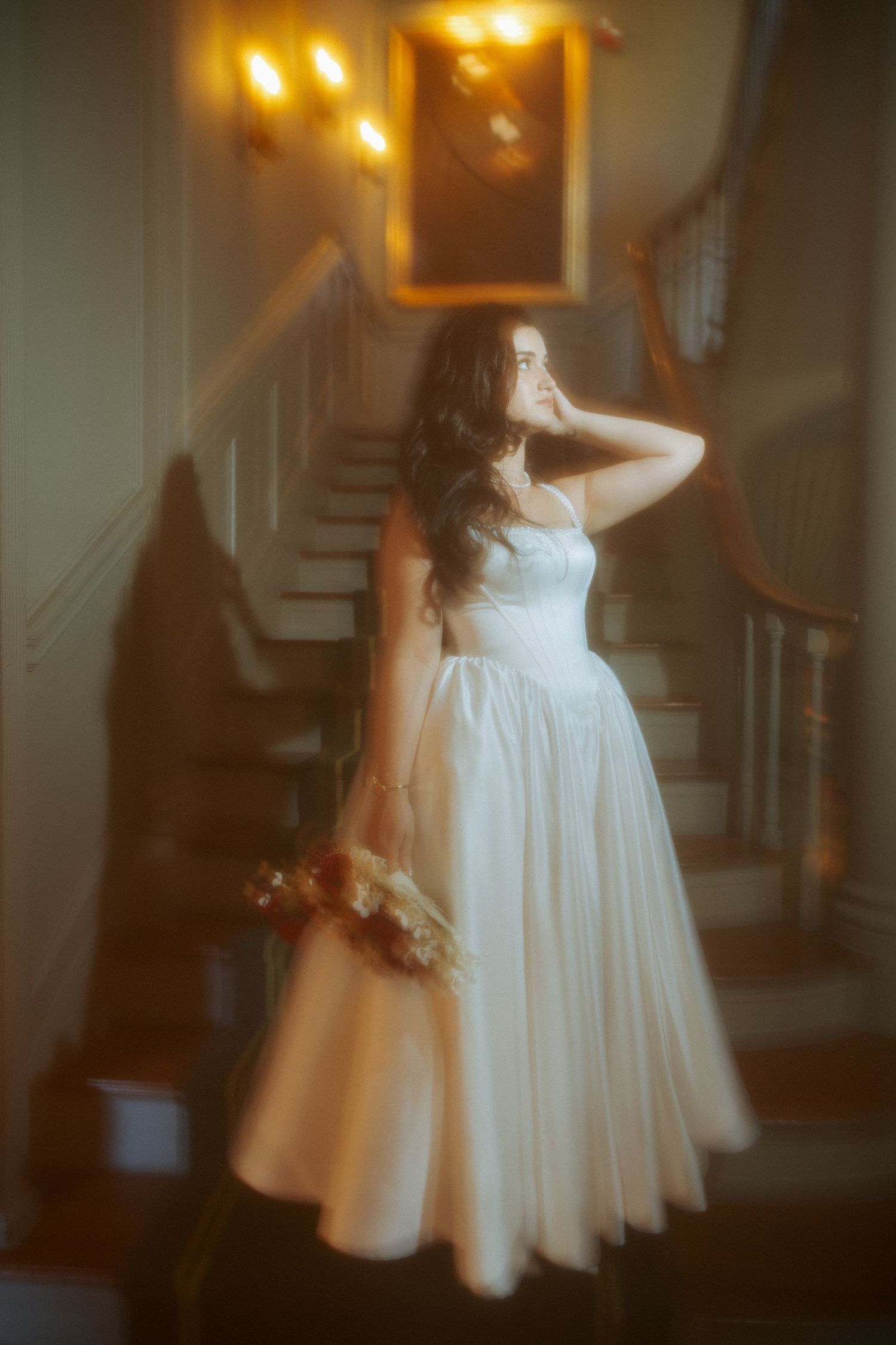 Bride in white gown holding bouquet stands by staircase in warm glowing light.