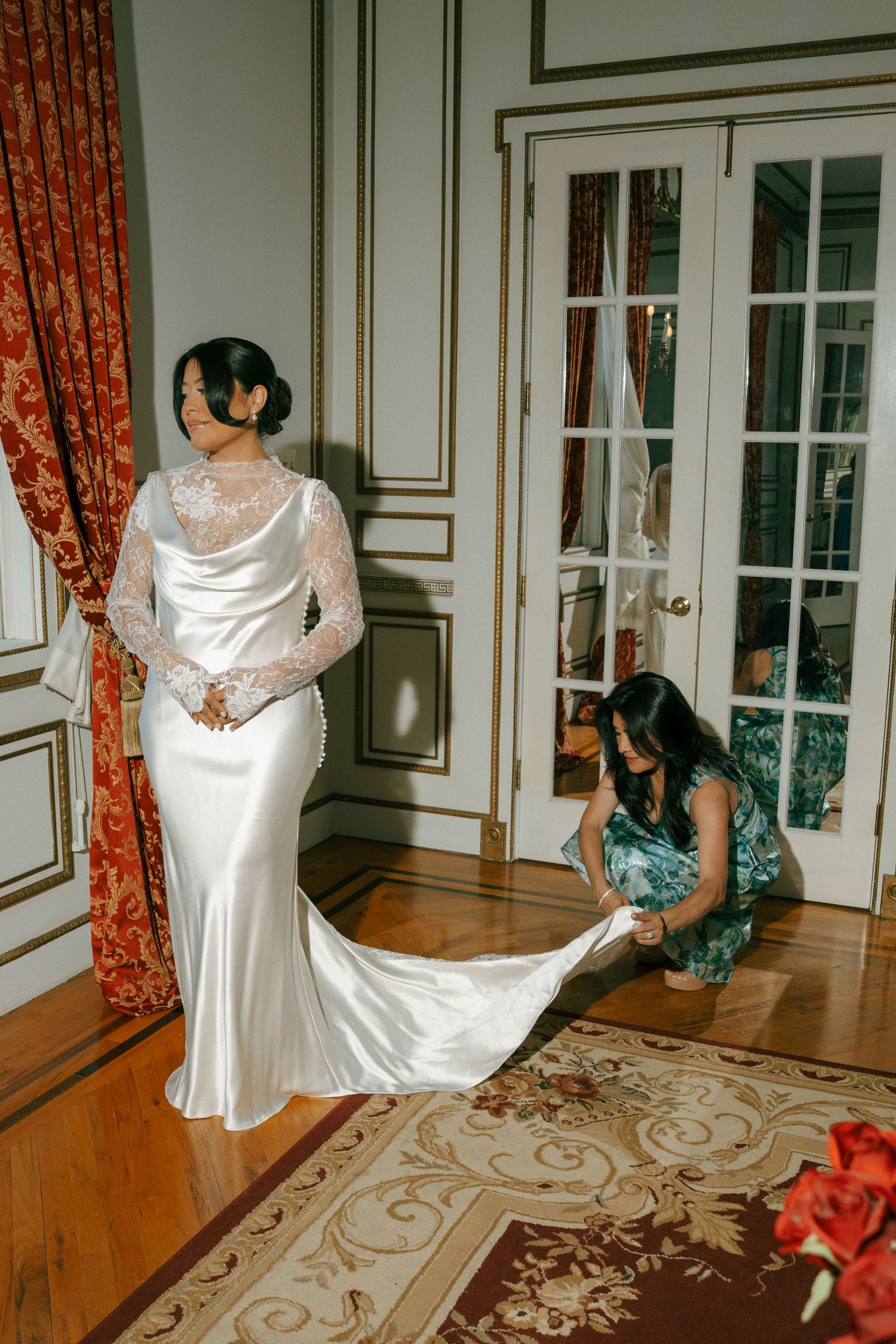 Bride in elegant white satin gown with lace sleeves as bridesmaid arranges train in ornate room.