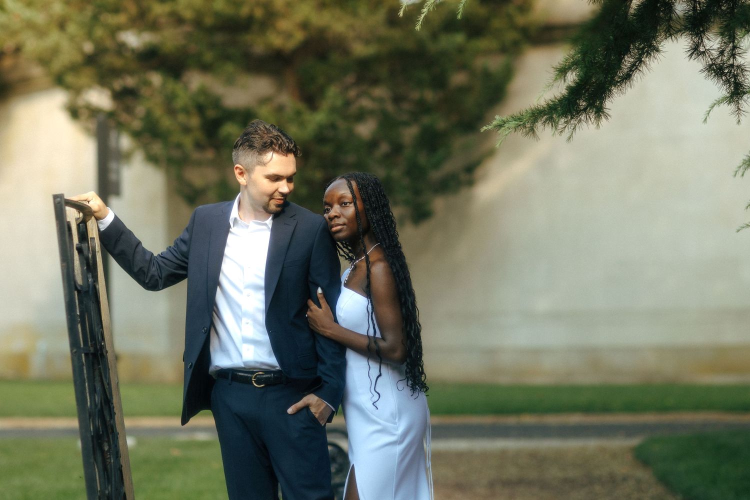 Interracial couple posing outdoors near a gate surrounded by lush green trees in casual formal attire.