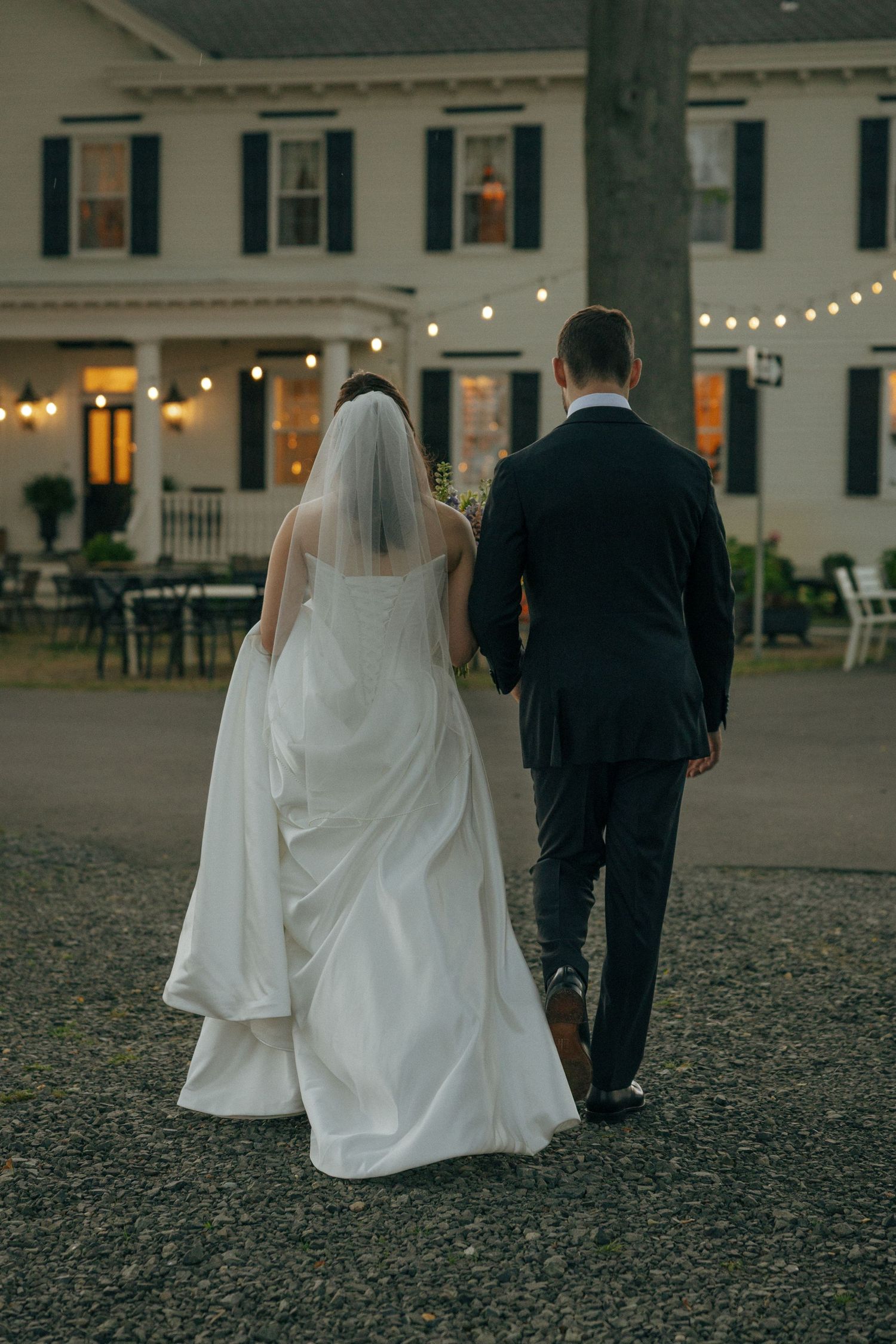 Bride and groom walk hand in hand toward an elegant venue adorned with string lights at dusk.