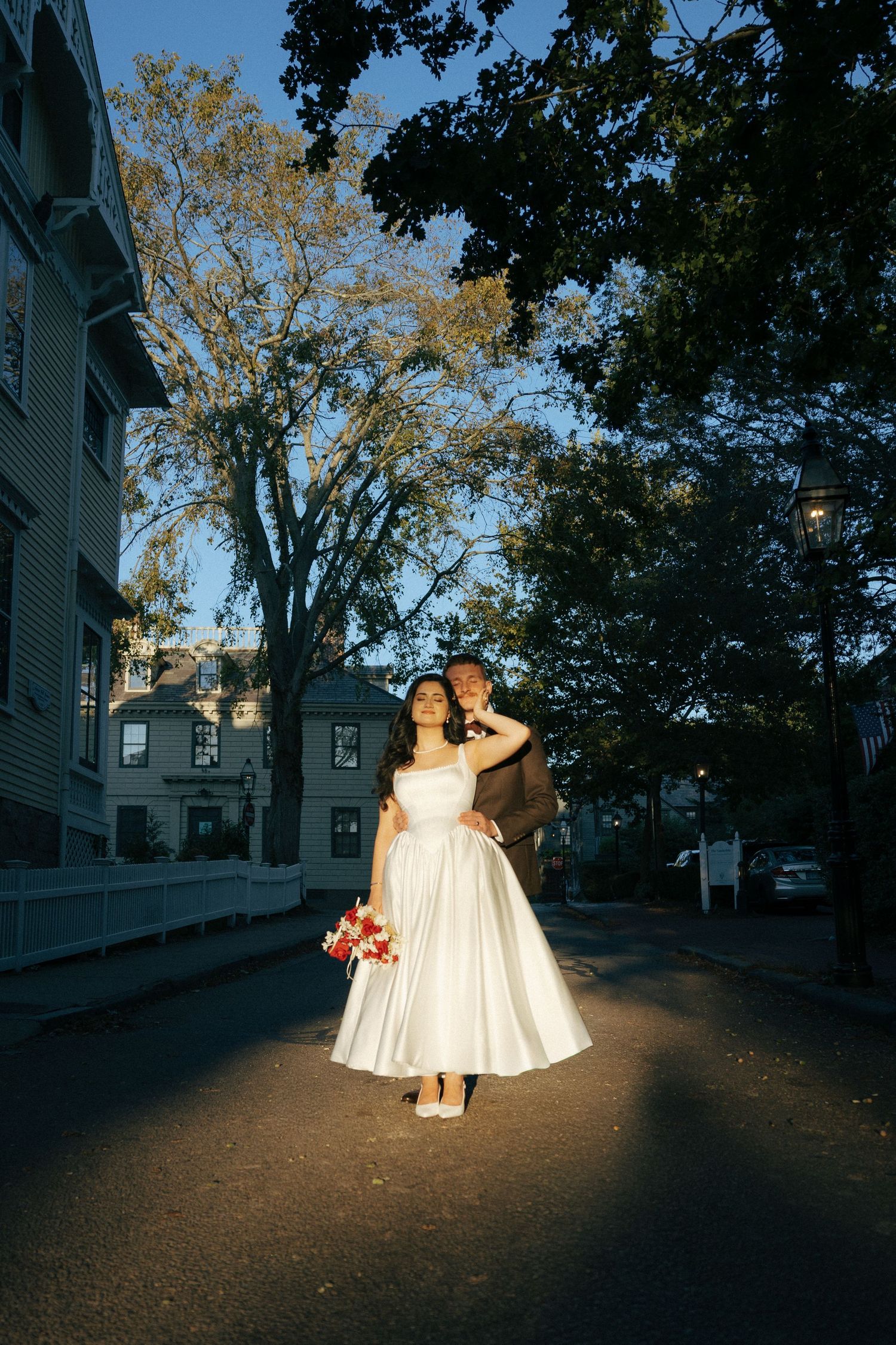 Bride and groom stand in sunlit street surrounded by tall trees in golden hour light.