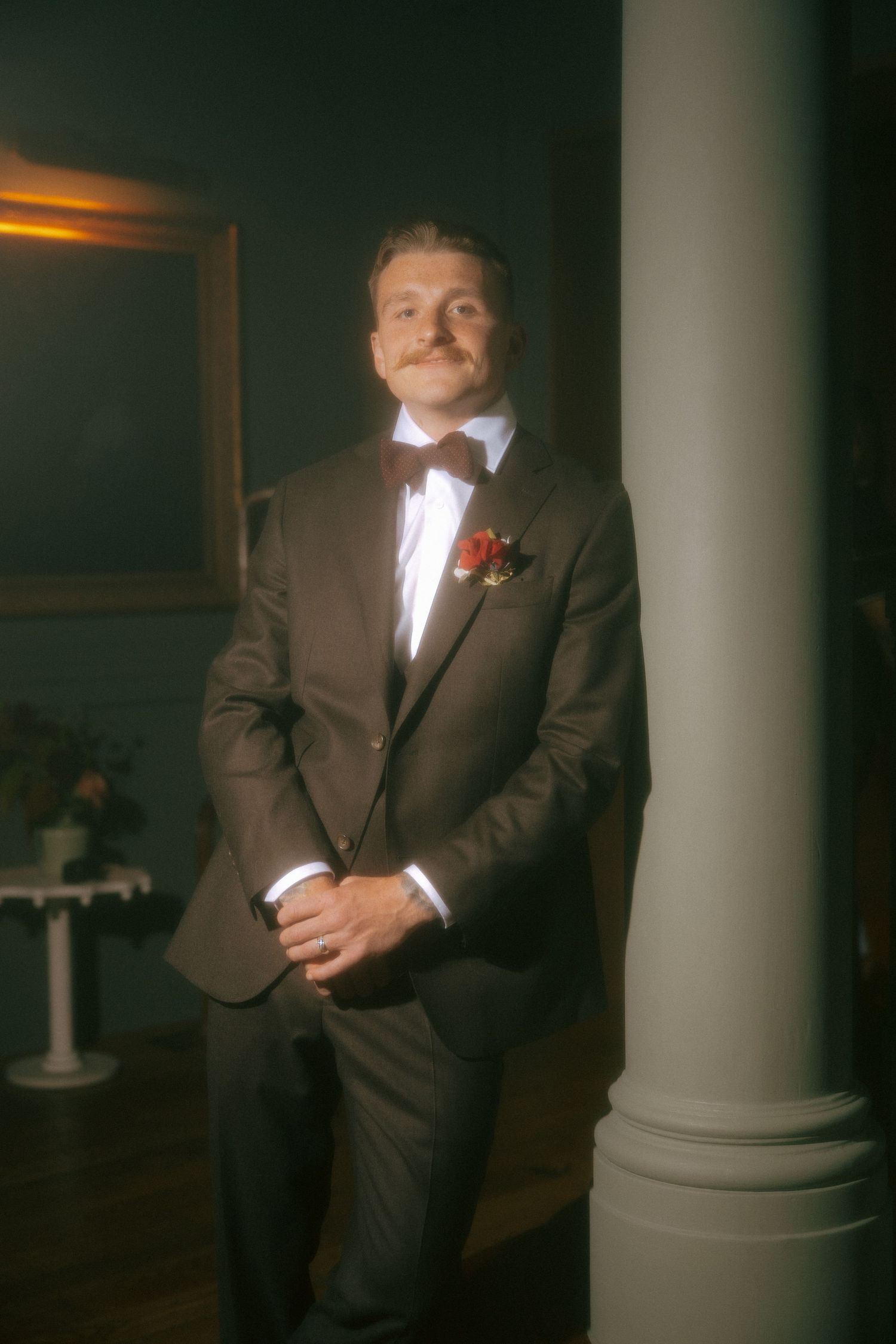 Dapper groom in brown suit with red boutonniere and bow tie leaning against column in elegant venue.