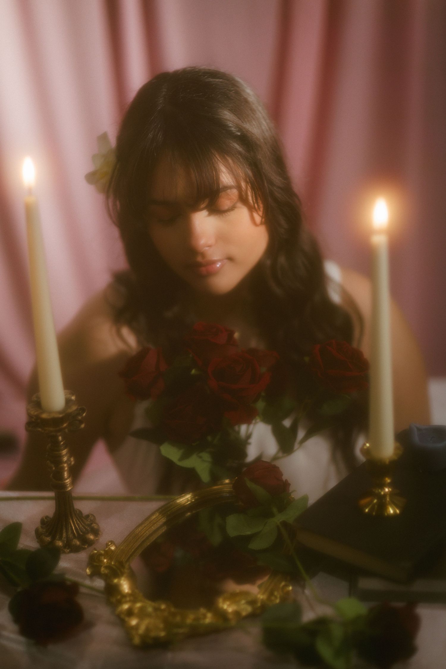 Woman with flower in hair gazing down at gold mirror on candlelit table with red roses.