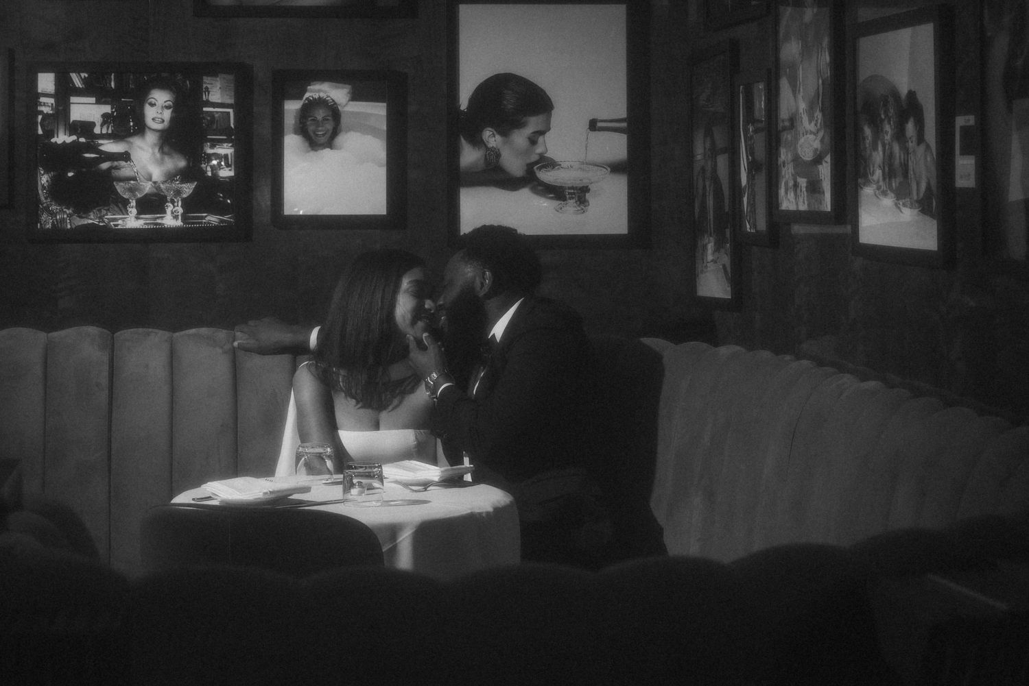 Black and white photo of a couple sharing an intimate moment at a restaurant booth, surrounded by framed photos on the wall.