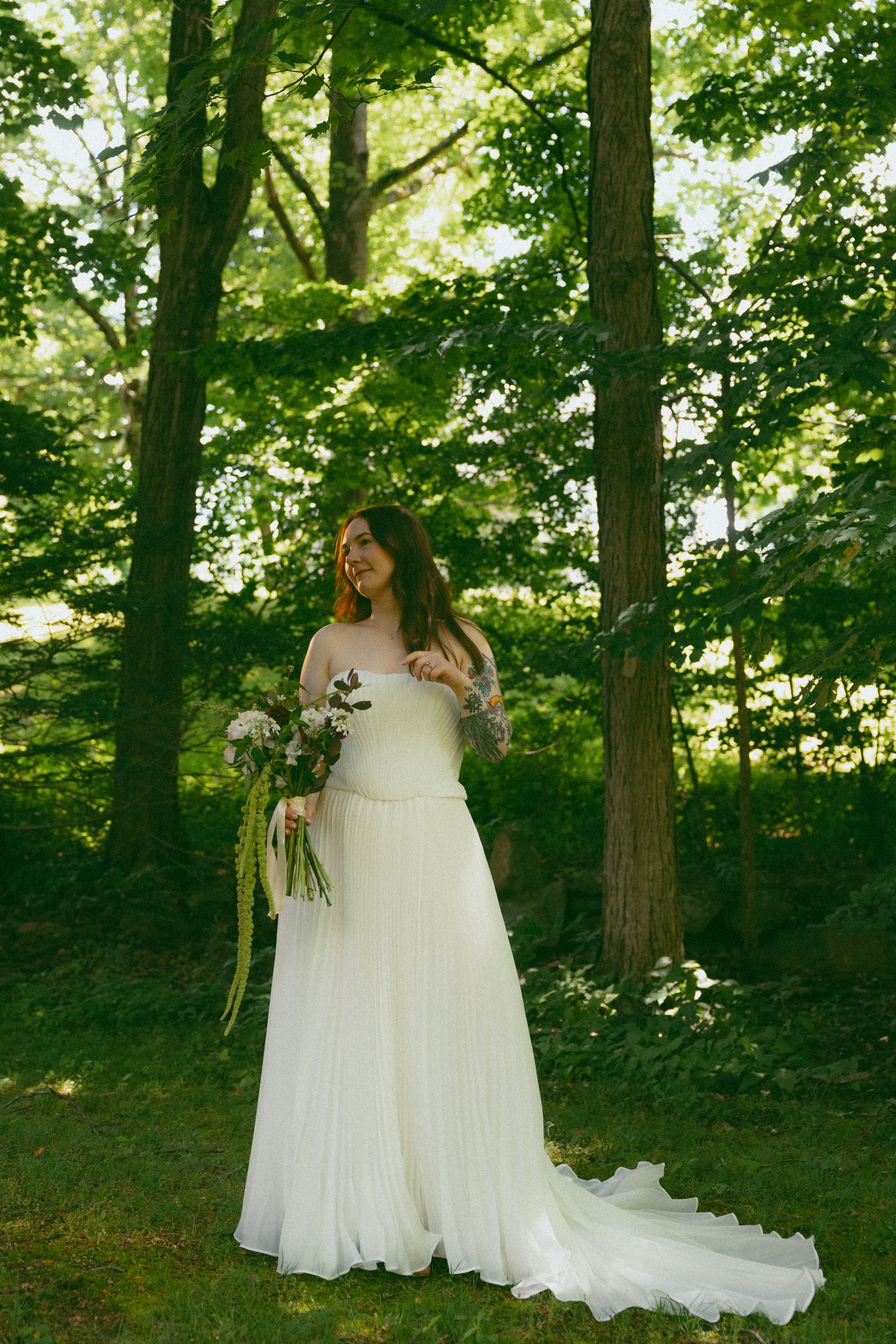 Bride in white strapless gown holding floral bouquet standing among lush green trees in an outdoor forest setting.