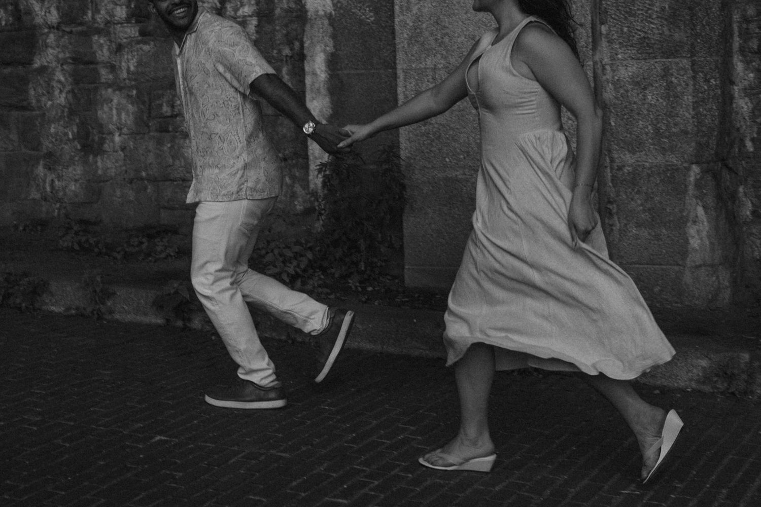 Black and white photo of a couple holding hands while running together along a stone wall.