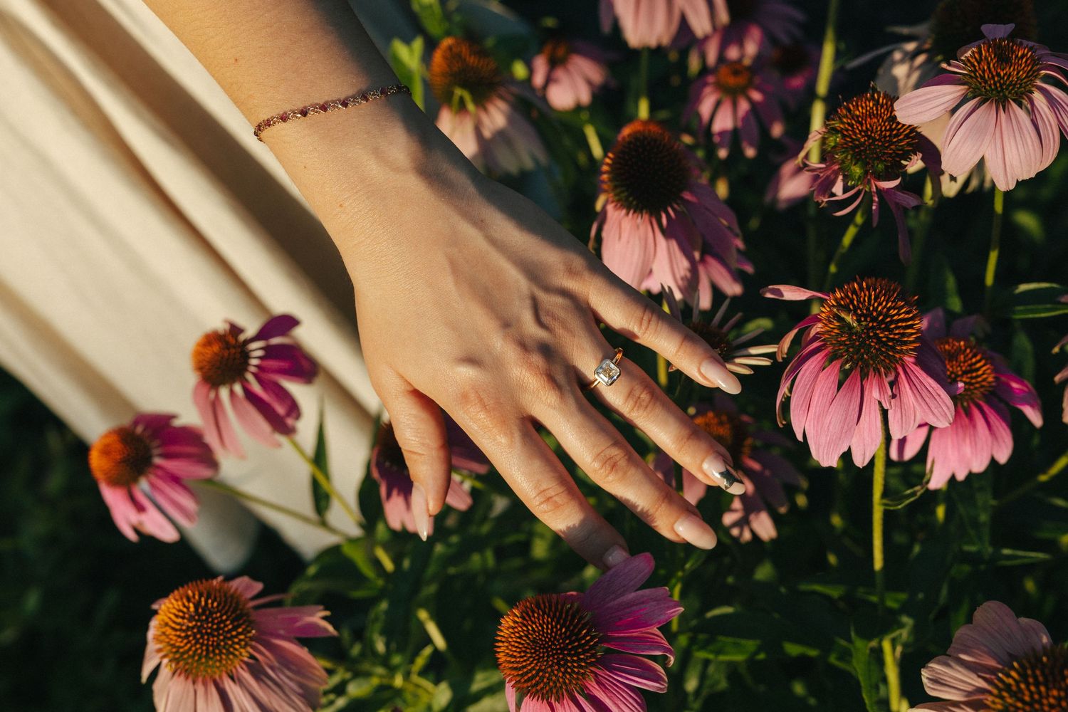 A hand with a delicate bracelet and ring gently touches pink echinacea flowers in a garden.