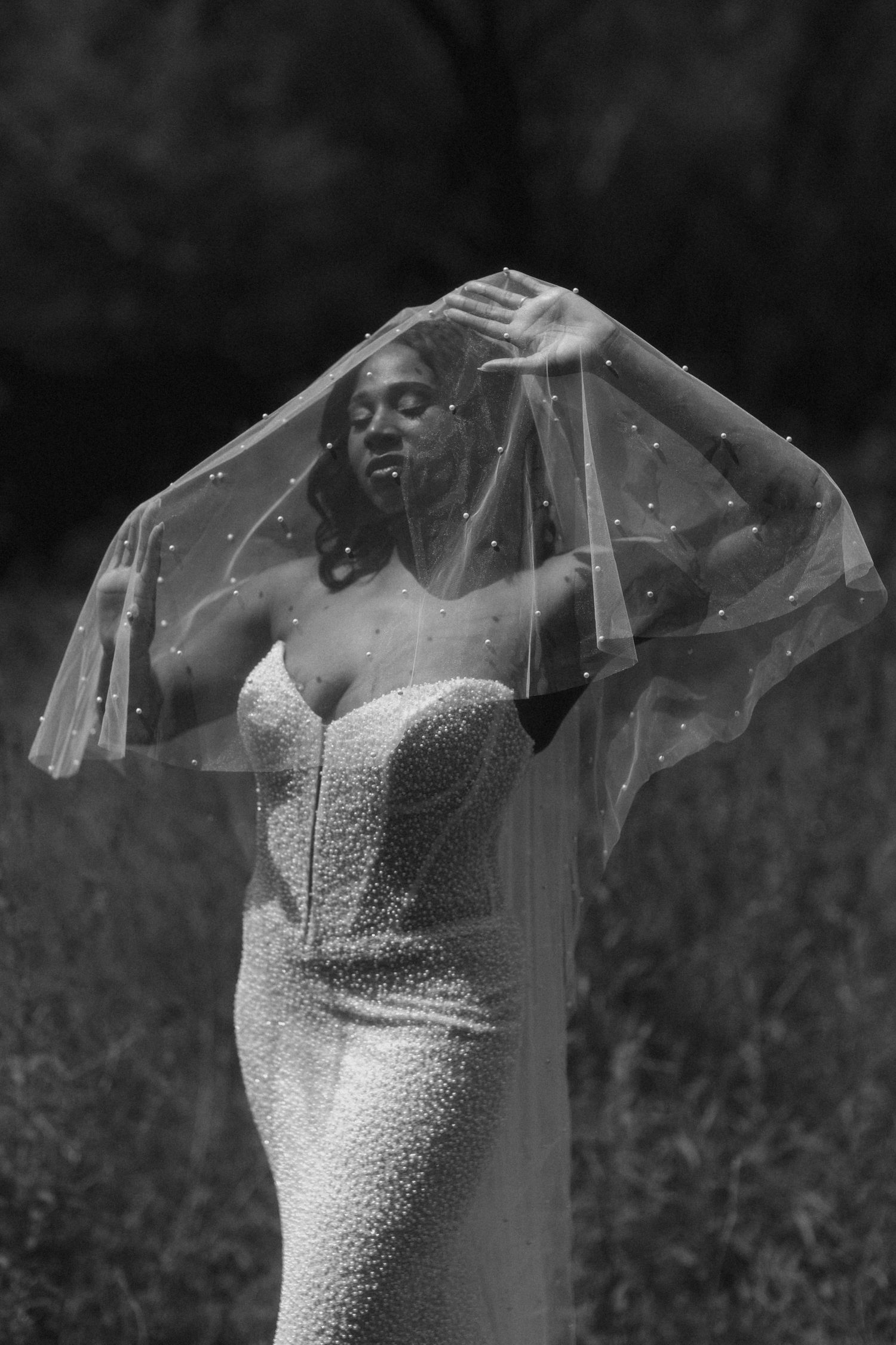 Black and white photo of a bride in a beaded gown lifting her pearl-embellished veil outdoors.