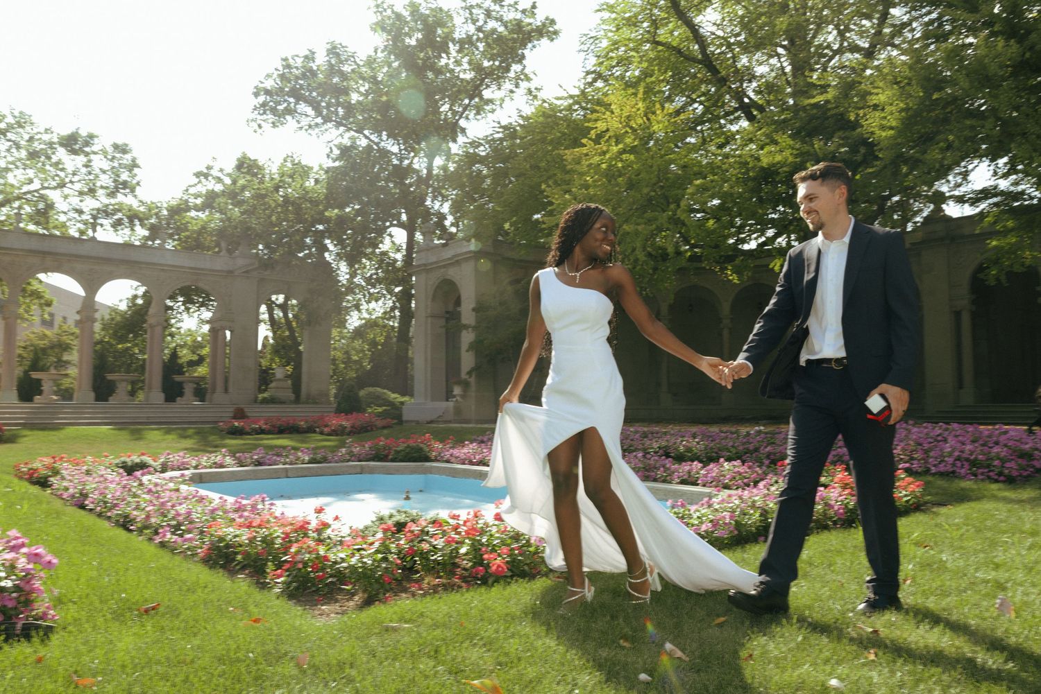 Bride and groom holding hands in elegant garden with fountain, colorful flowers, and stone columns backdrop.