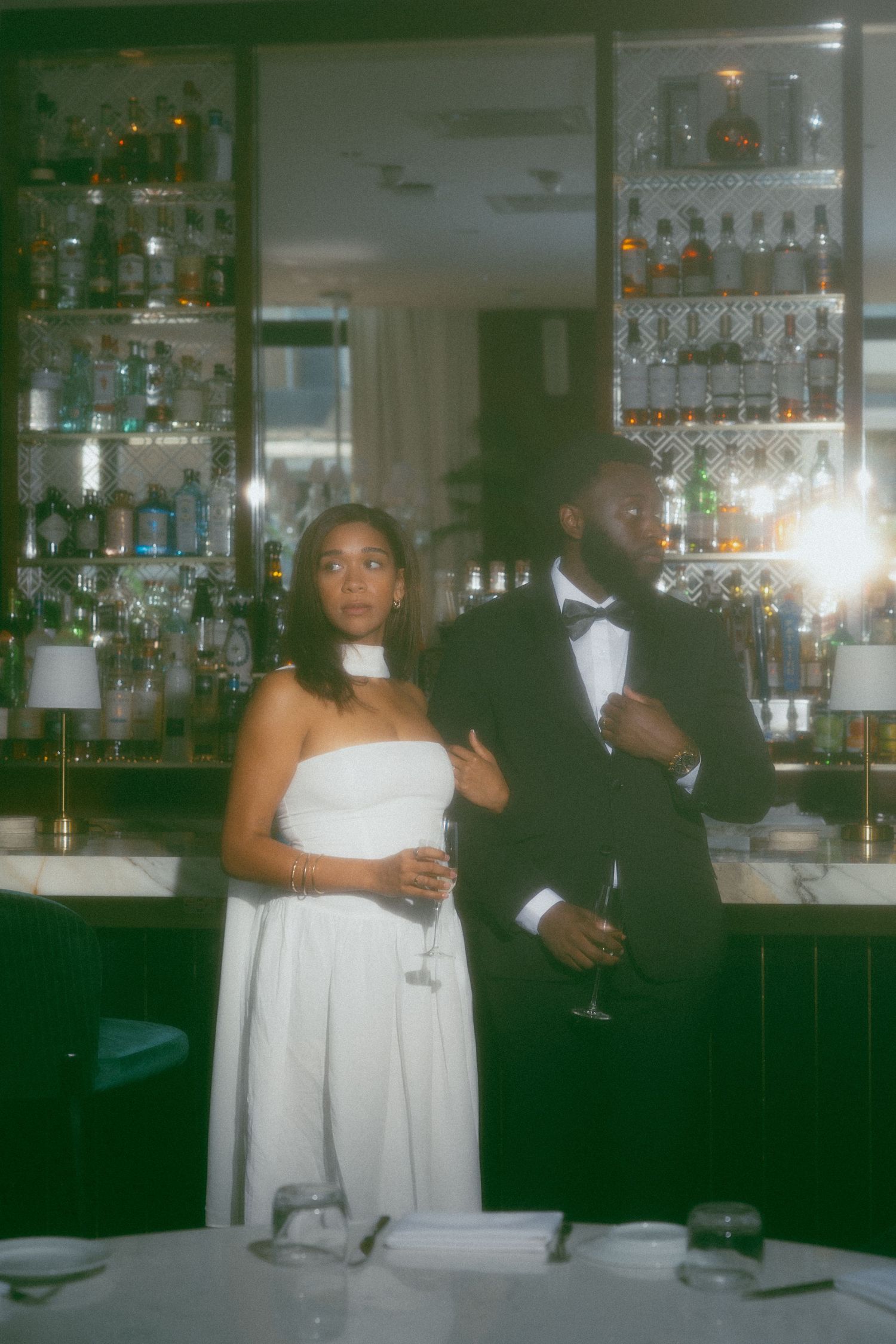 Bride in white strapless gown and groom in black tuxedo standing together at an elegant bar venue.