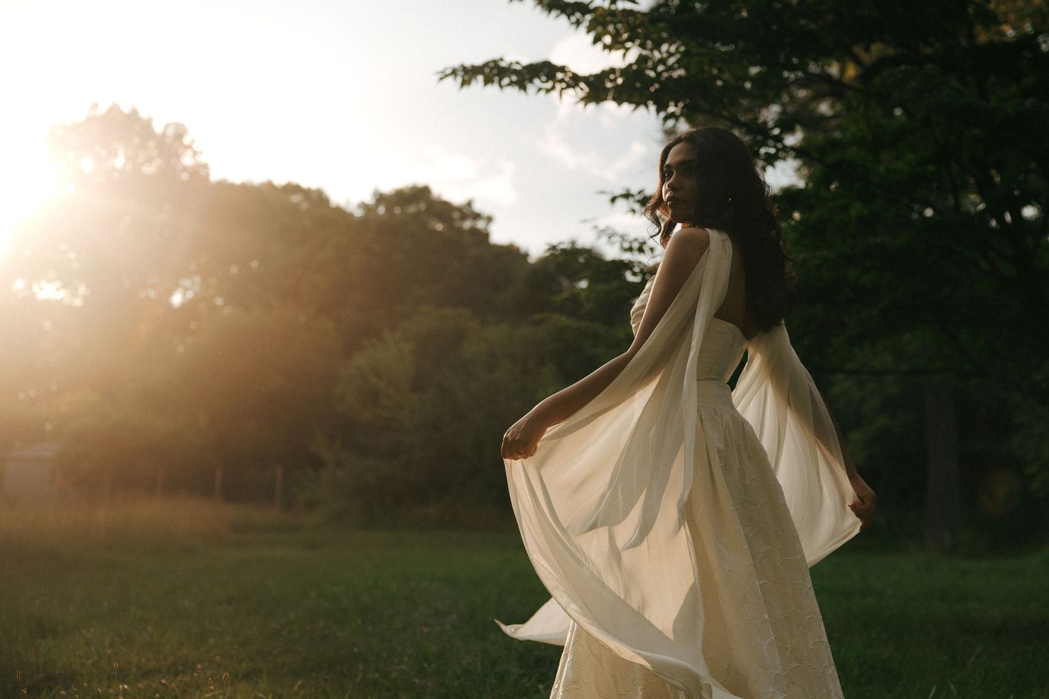 Woman in flowing white dress twirling in sunlit park at golden hour with lush green trees in background.