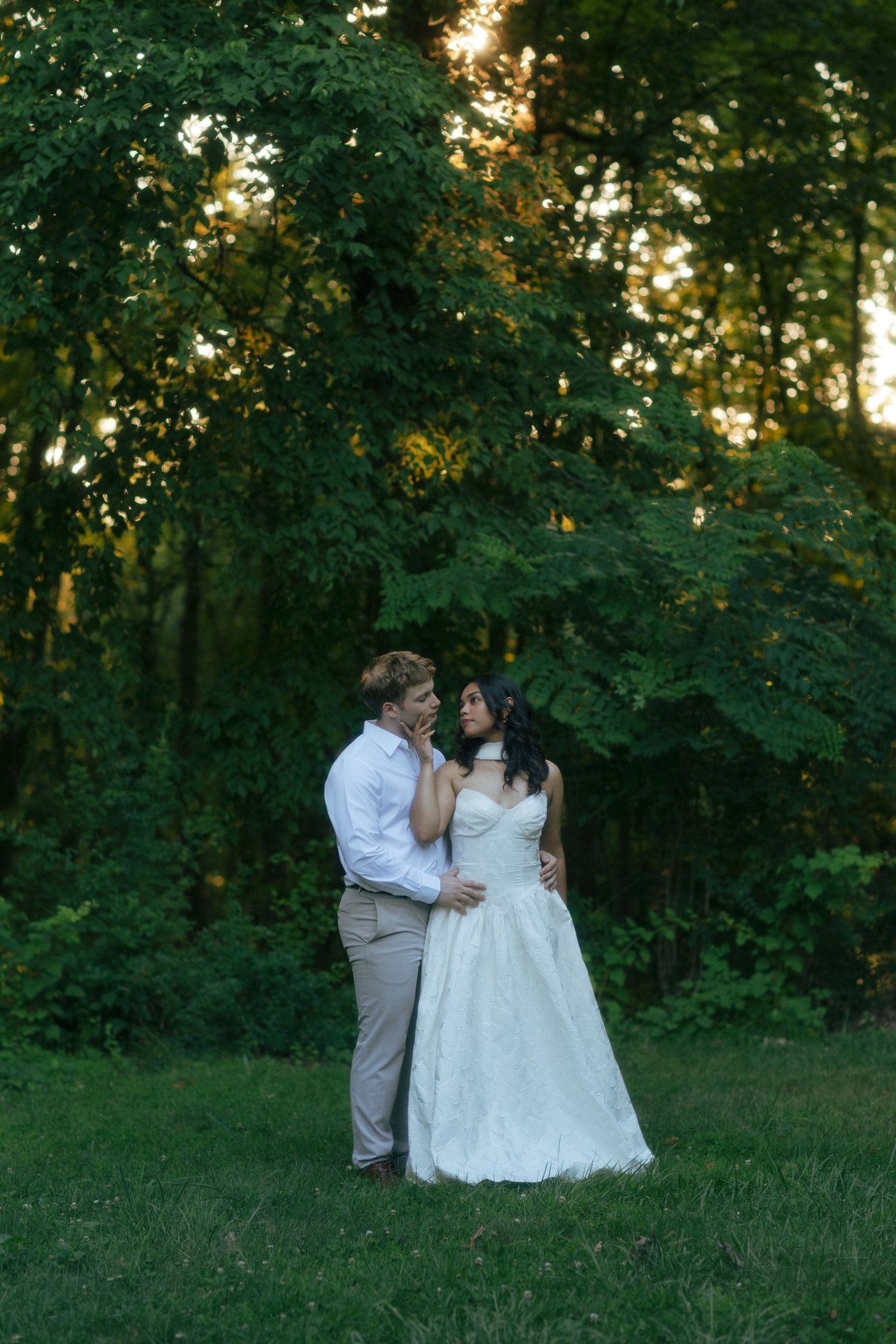 Bride and groom share a romantic moment in a lush green forest at golden hour sunset.