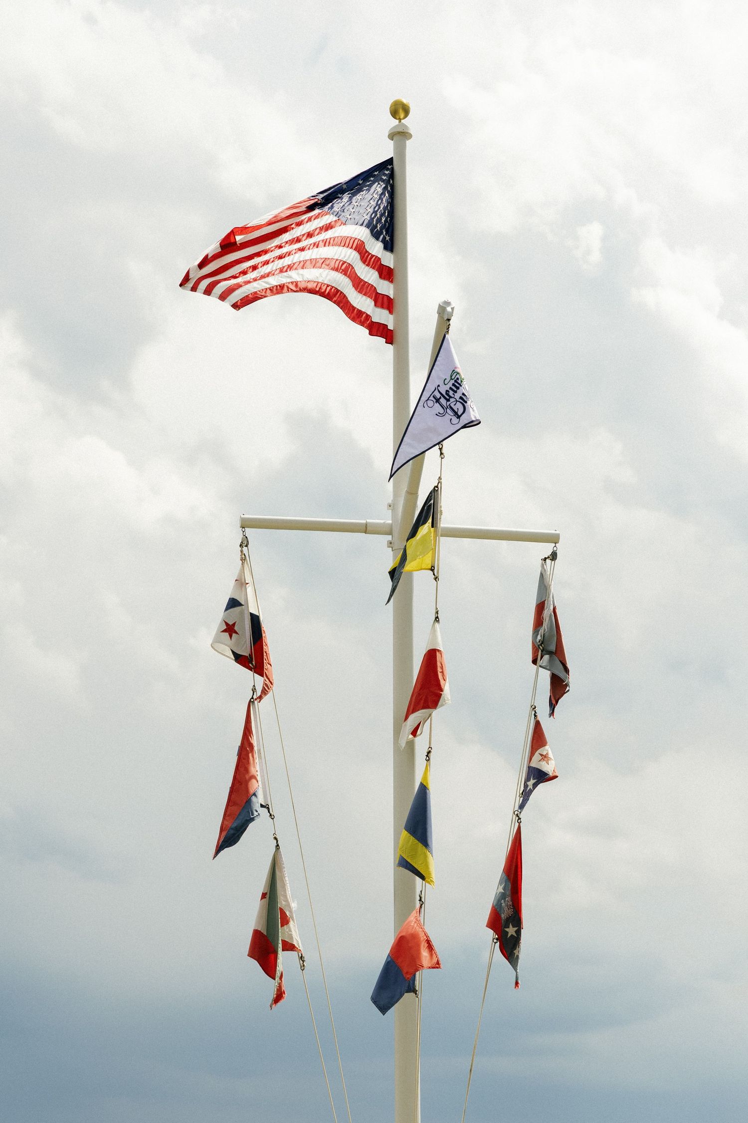 Multiple flags wave against a cloudy sky including an American flag and other colorful banners.