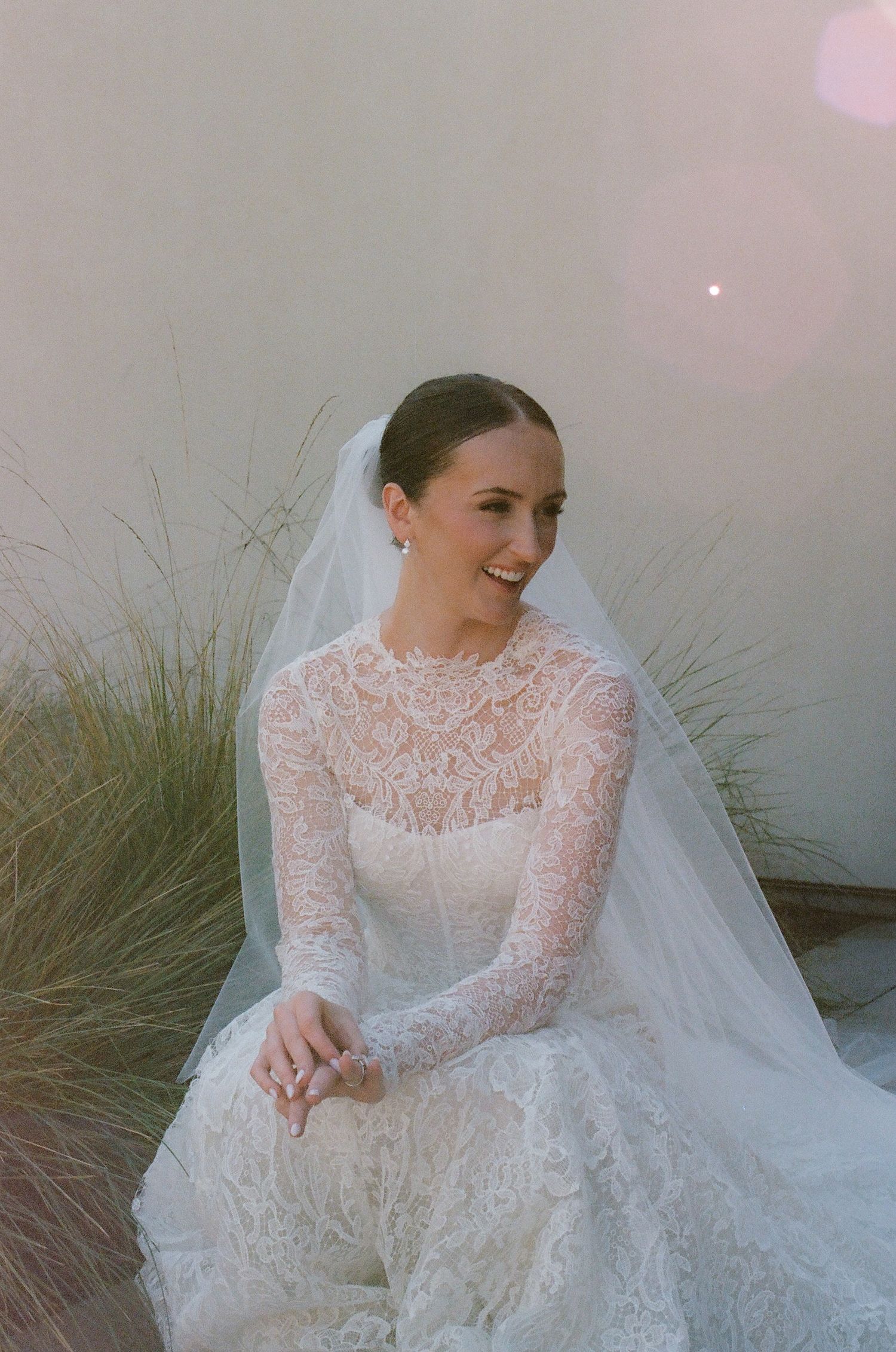 Bride in long-sleeved lace gown poses gracefully against natural backdrop.