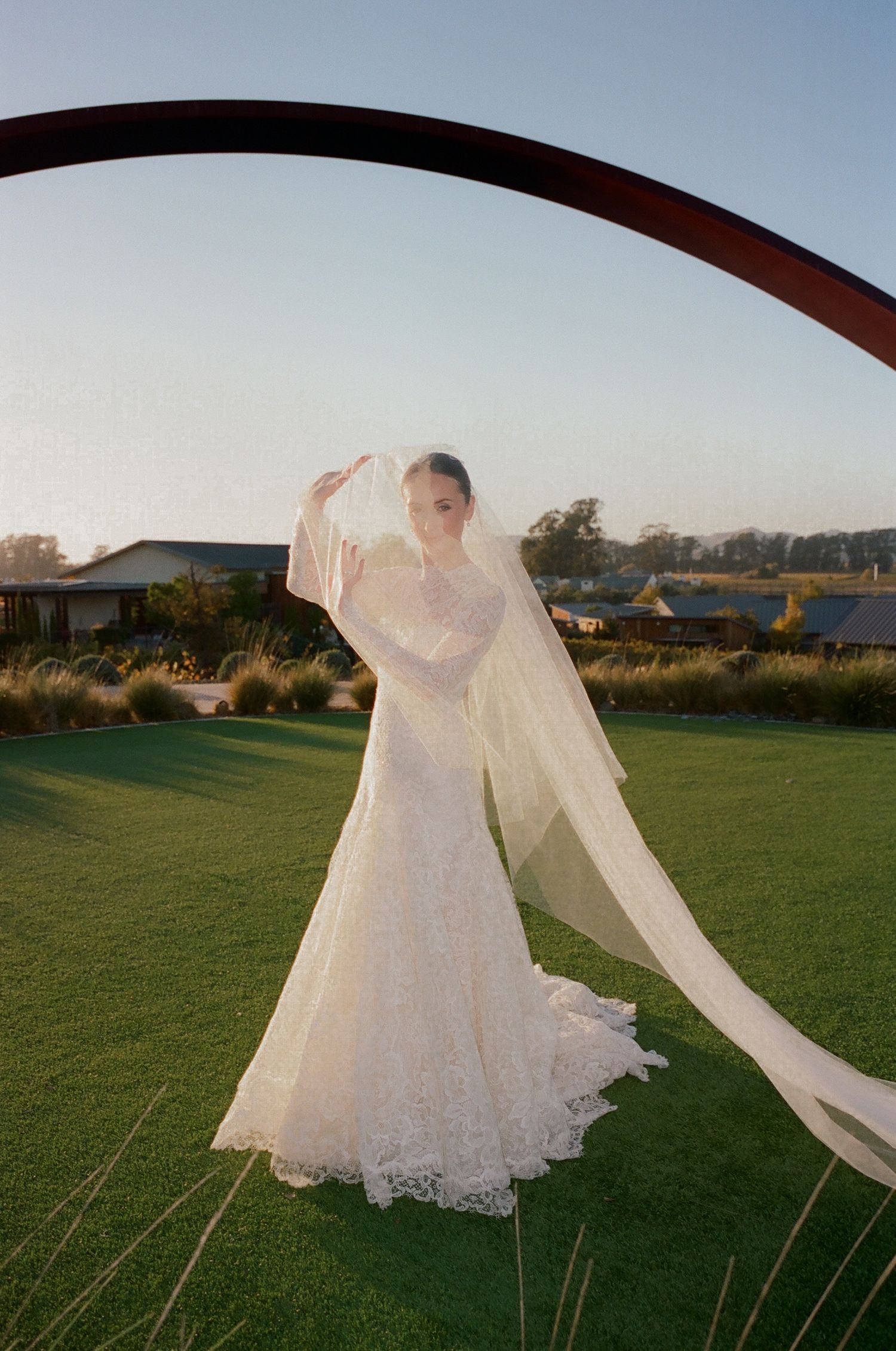 A flowing wedding dress and veil catch the sunlight beneath a curved metal arch.