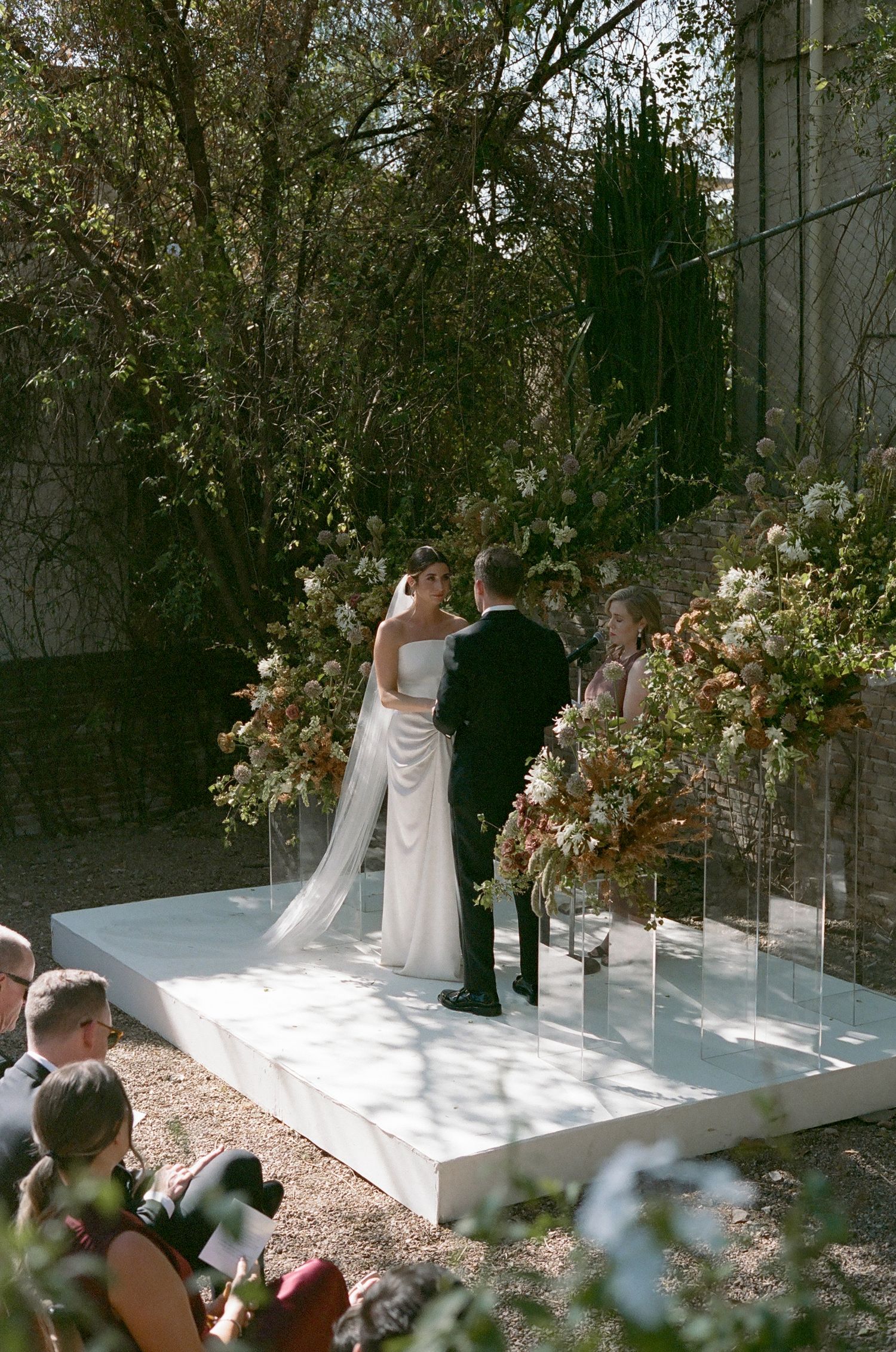 Outdoor wedding ceremony with floral arrangements on white platform surrounded by trees.