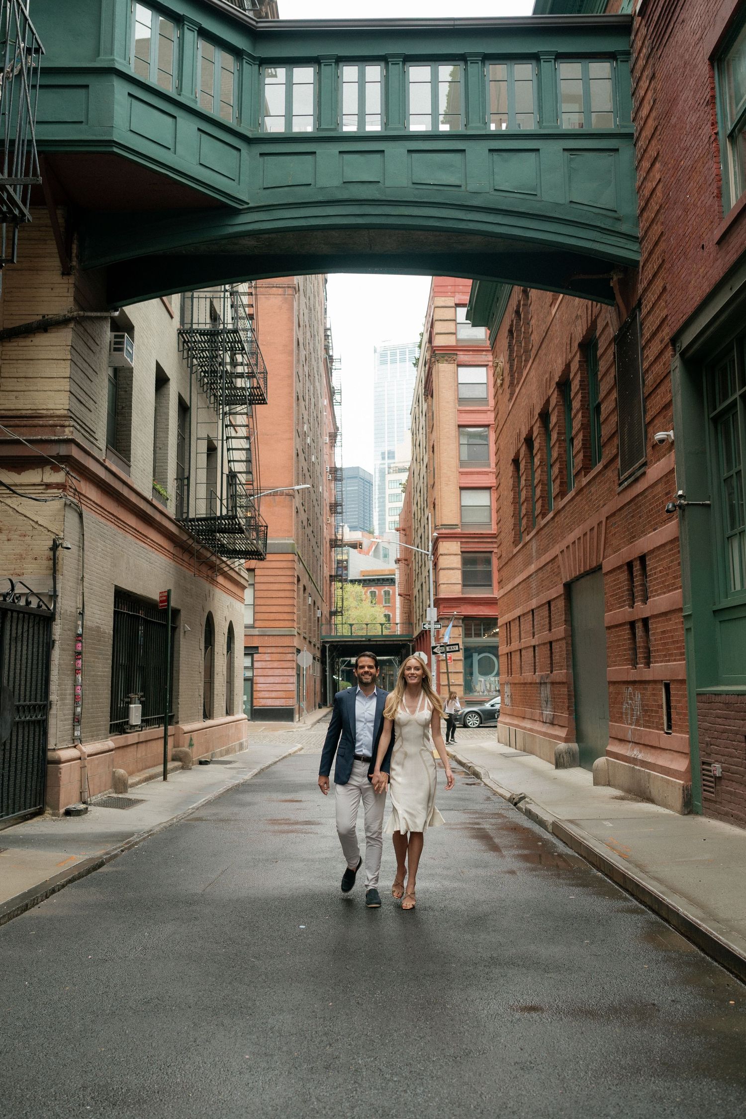 A couple walks down a historic cobblestone street with green archways overhead.