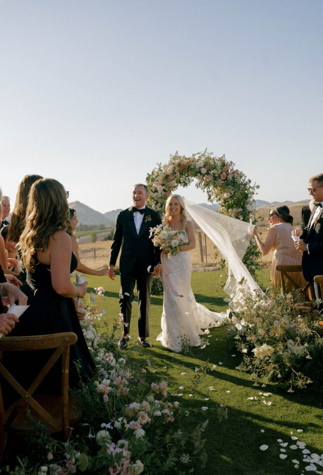 Outdoor wedding ceremony with floral arrangements and guests on a hillside at sunset.