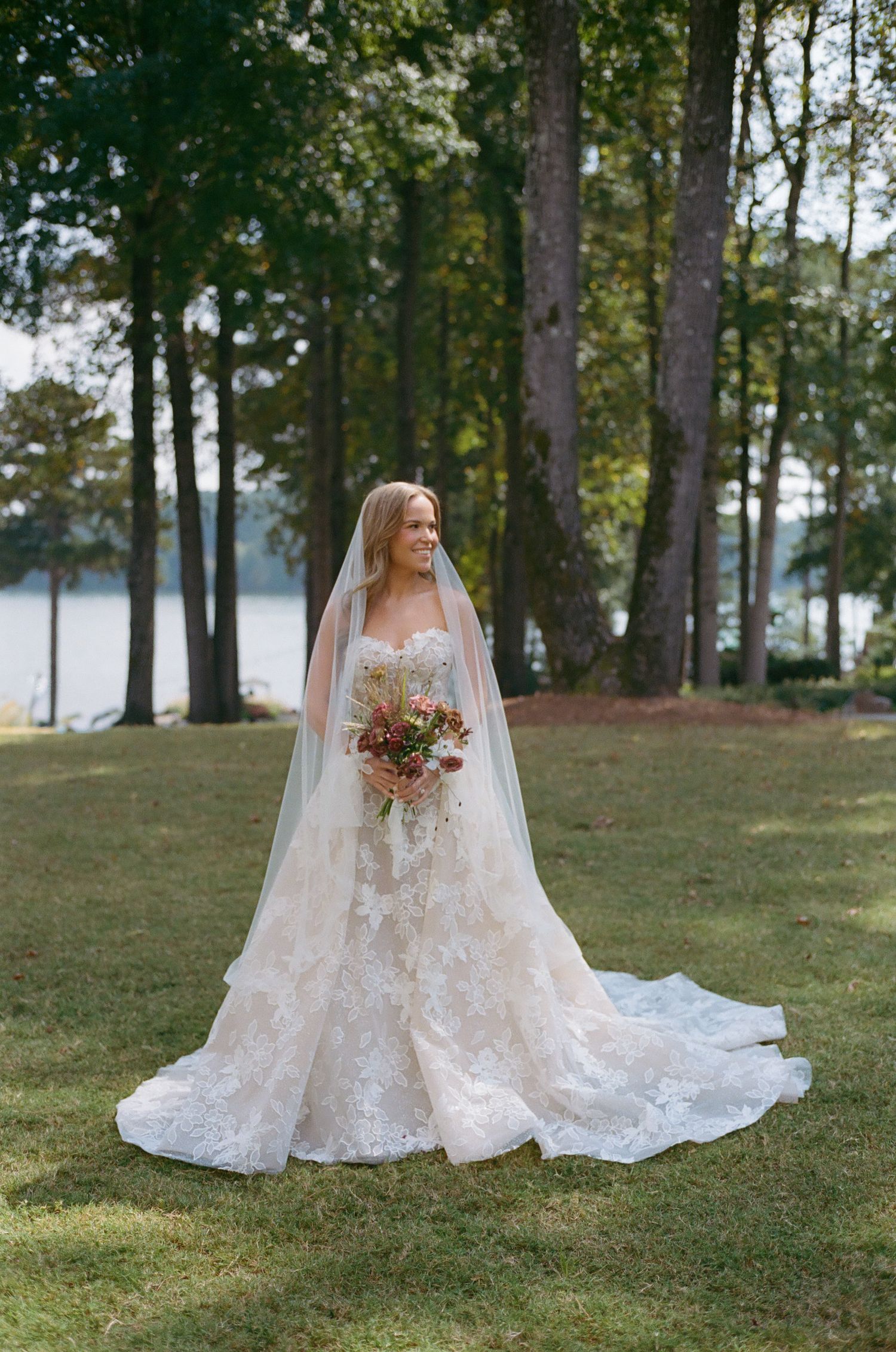 Stunning bridal portrait in lace wedding dress against lakeside forest setting.