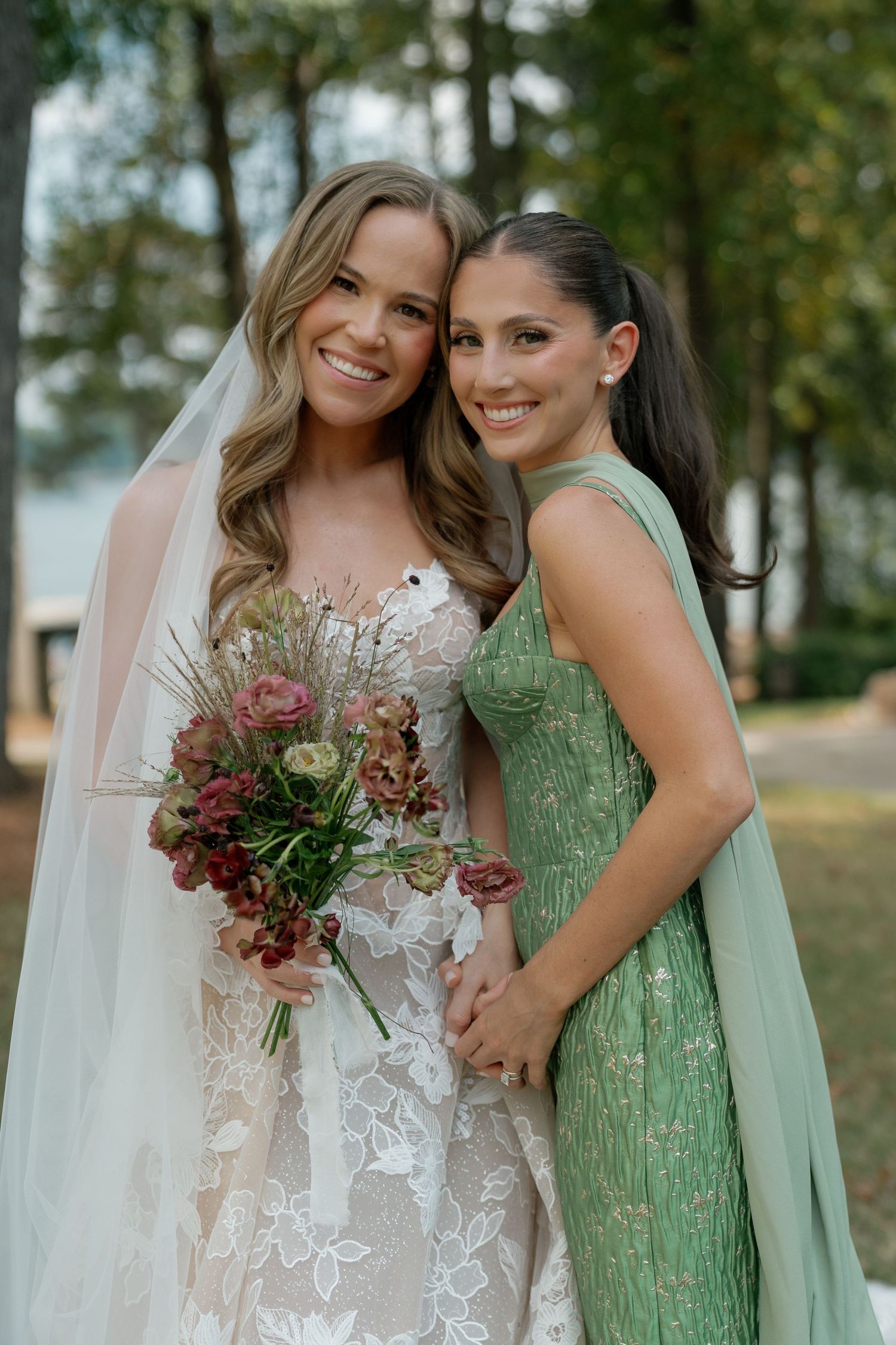 A bride in a lace gown poses with a bridesmaid in a mint green dress holding a pink bouquet.