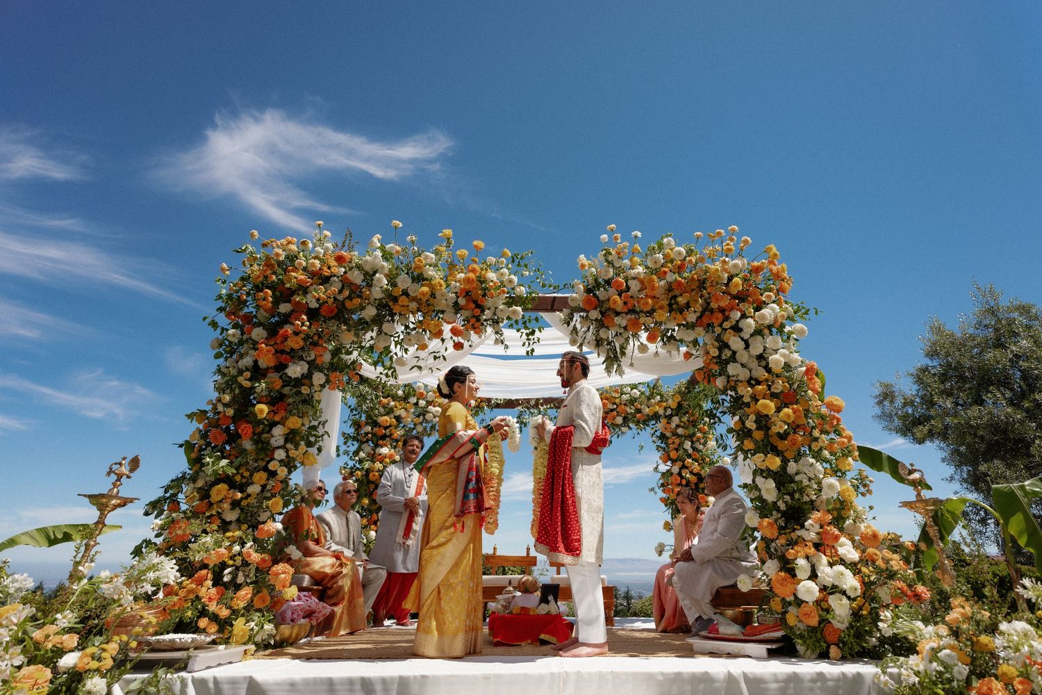 Indian wedding ceremony under floral arch with orange and white blooms against a blue sky.