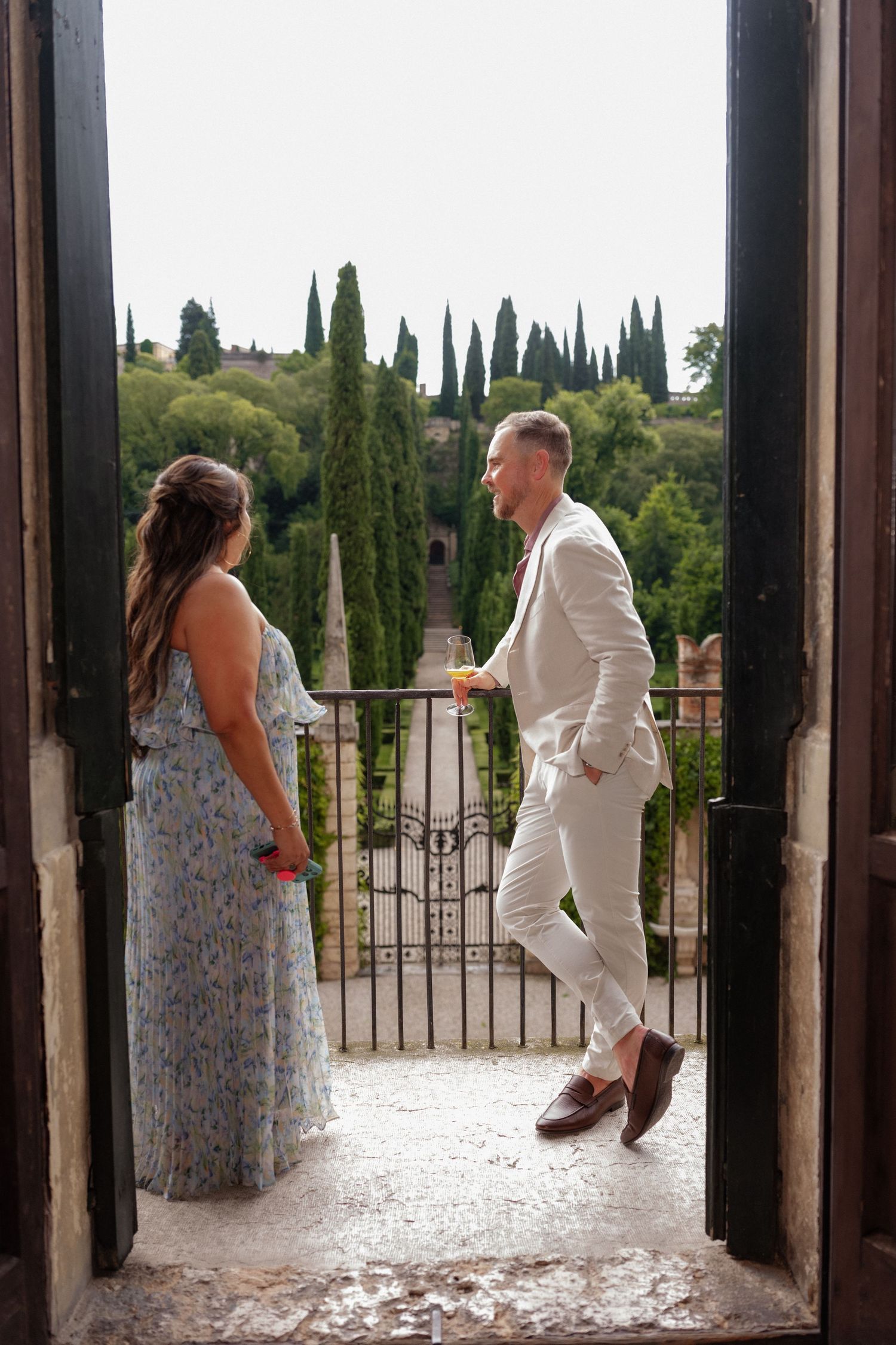 Couple in elegant attire standing on a balcony overlooking lush Italian gardens with cypress trees.