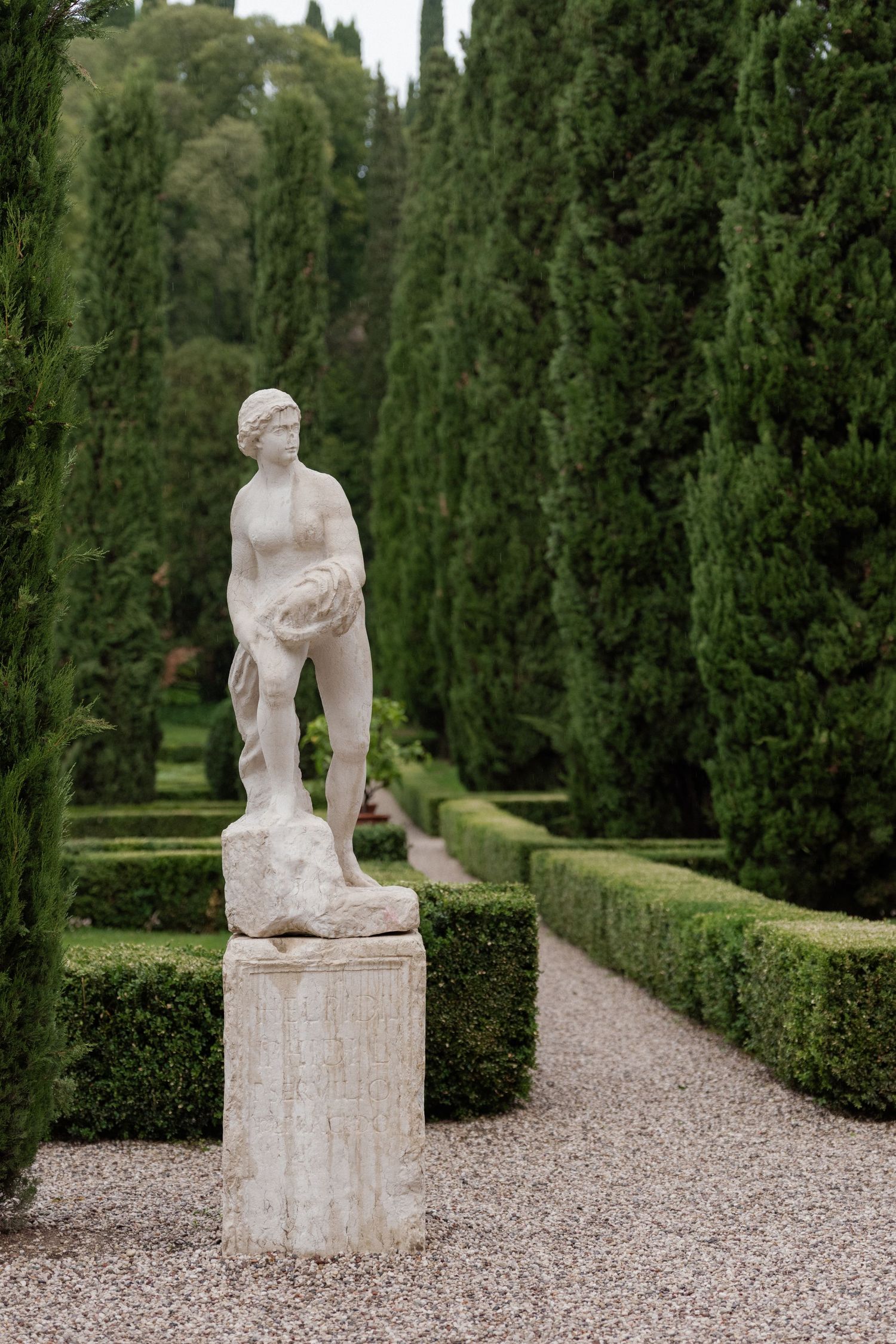 Classical marble statue of a female figure on a pedestal in a formal Italian garden with cypress trees.
